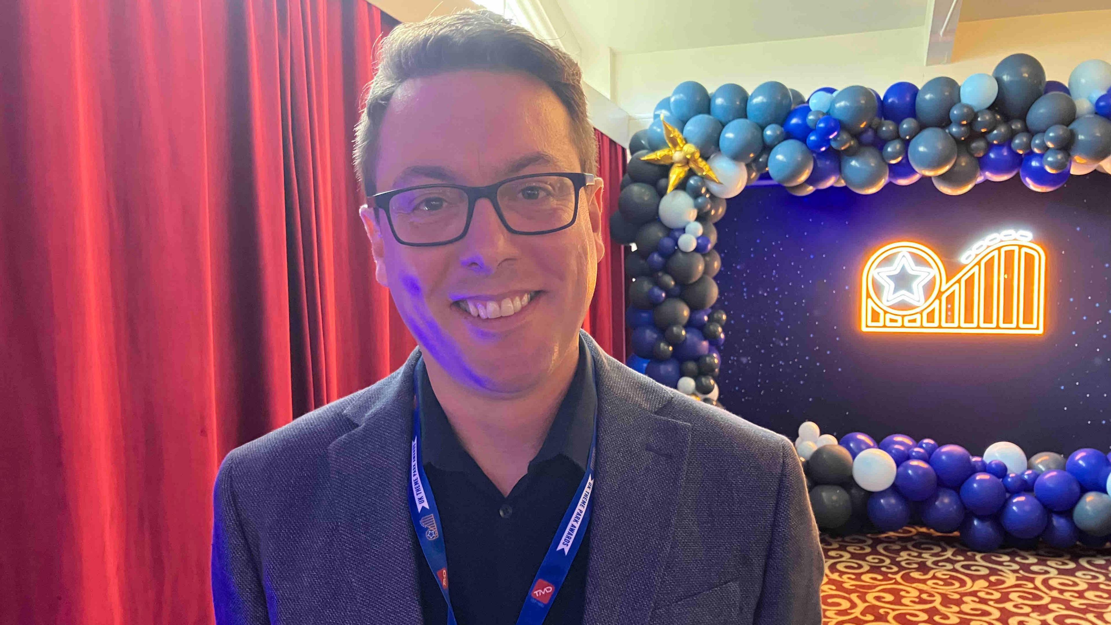 Man standing in front of a blue and white balloon-decorated backdrop with an illuminated sign featuring a star and roller coaster design.