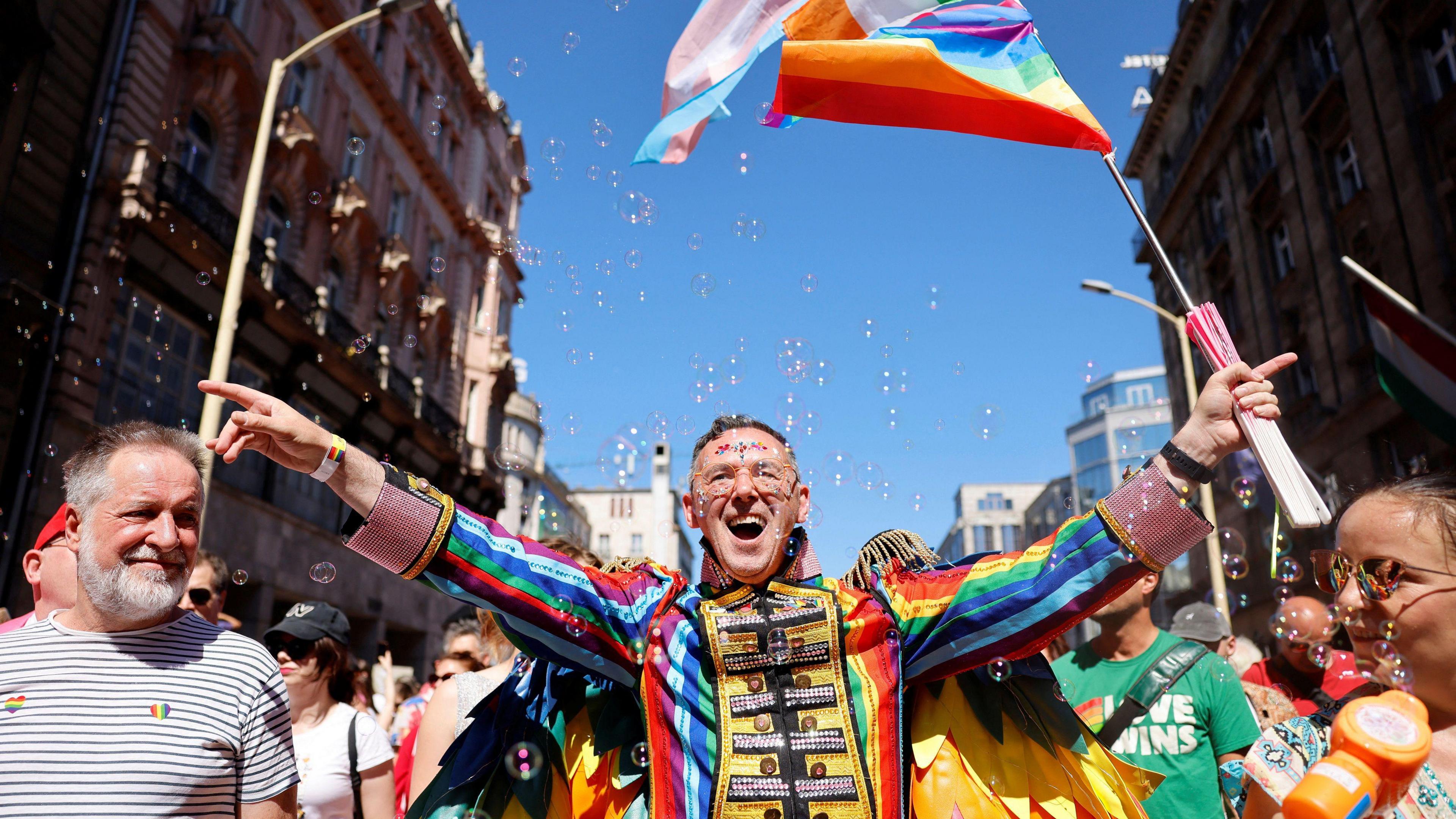 A man standing in the march wearing a multicoloured ringmaster type outfit. He has both his arms out and is holding a pride flag in his left hand. He is smiling and wearing glasses. To his left - in the bottom right corner of the picture - is a woman in sunglasses with a bubbles gun - there are bubbles across the scene. On his right, there is a smiling man in a white-and-black horizontally-striped t-shirt. He has two hear pride stickers on it.