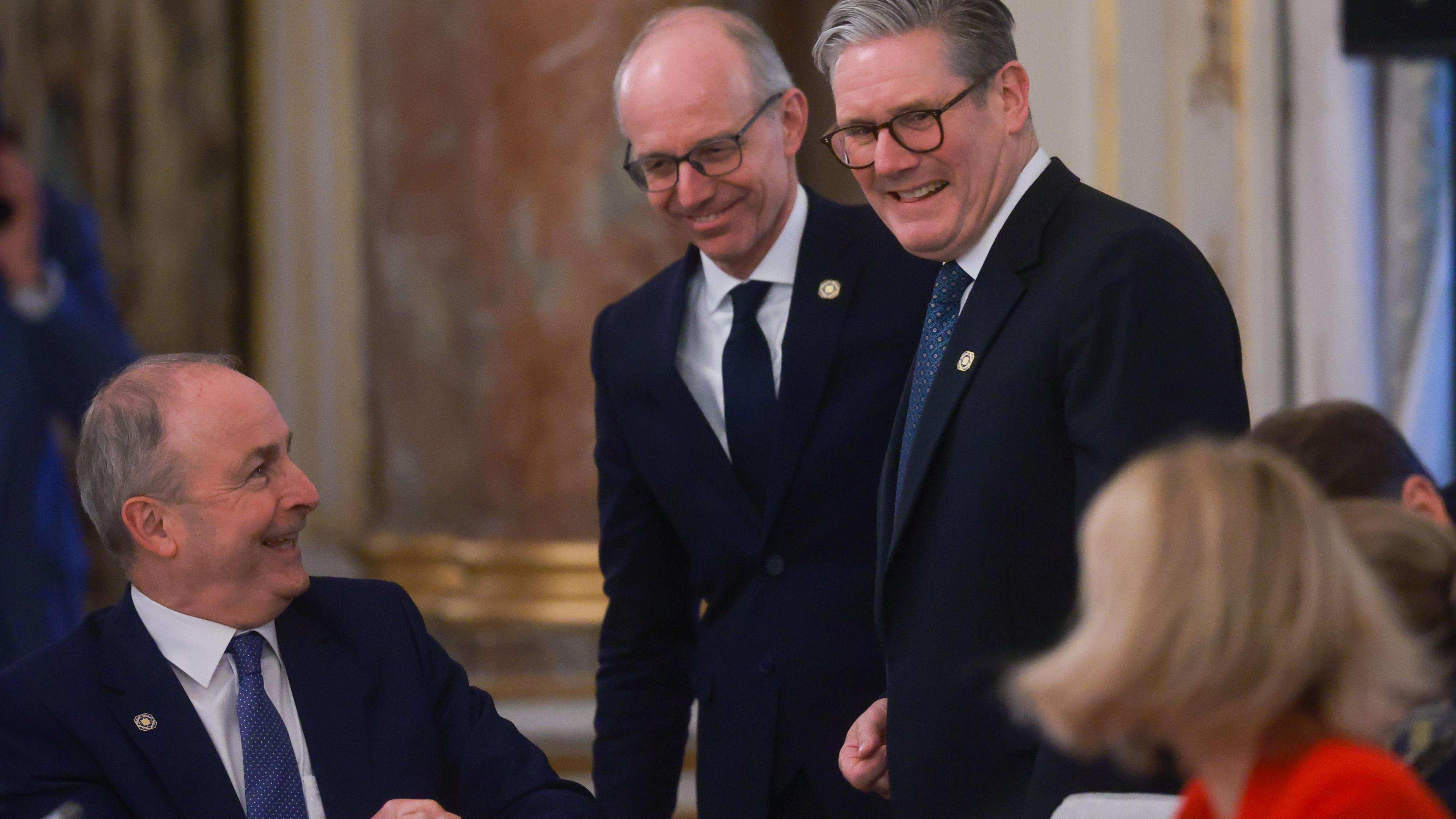 Left to right: 
Micheál Martin is smiling from a seated position at Keir Starmer who is standing and smiling at a woman wearing red who is seated to his left with blond hair to her shoulders with her face turned towards him and away from the camera. Behind Keir, standing is Luc Frieden who is also smiling. Behind the four of them, is a regal looking gold frame with an autumnal coloured pastel painting. 
Micheál Martin is wearing a navy suit, white shirt and blue tie. He has short dark grey hair. 
Luc Frieden is wearing a navy suit, white shirt and navy tie. He has a bald head with short hair on either side and is wearing black square glasses. 
Keir Starmer is wearing a navy suit, white shirt and teal blue tie. He has grey hair brushed to the right and black glasses.
All three of the men are wearing a yellow badge on their suit jackets. 