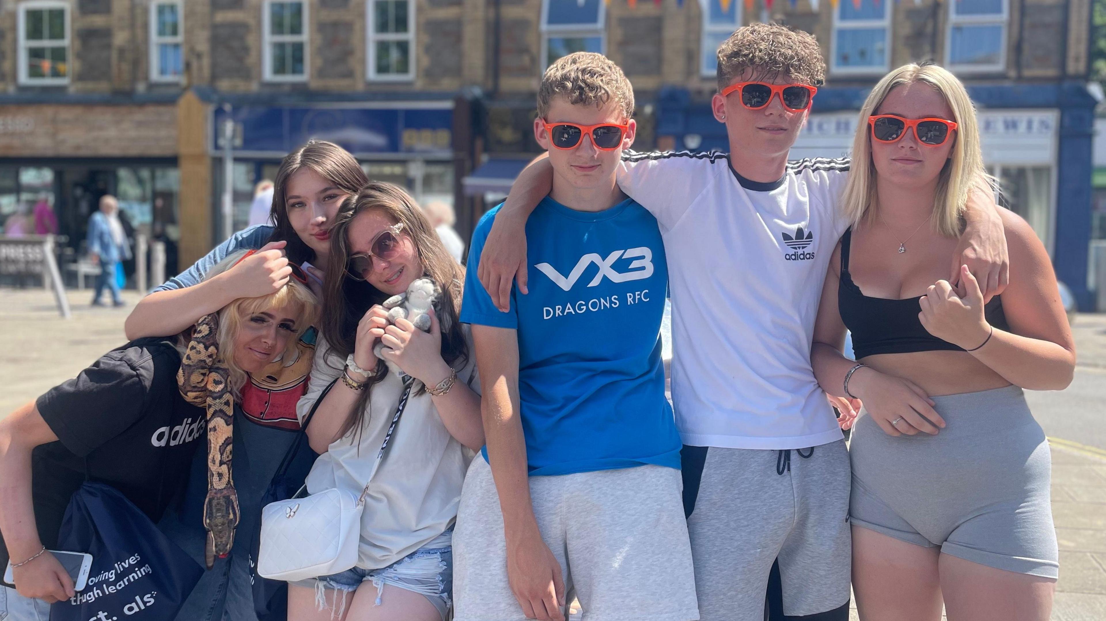 Six young people huddled together, divided into a trio of girls and two boys and a girl who are wearing orange-rimmed sunglasses. The girl on the right has blonde hair, grey shorts and a back crop top. The boy next to her has his arm around her, and has short curly light-brown hair and a white Adidas t-shirt on and grey shorts. His other arm is around a boy with short light-brown hair and a blue t-shirt which has VX3 Dragons RTC in white writing and grey shorts. Next to him are three girls huddled together. The one closest to him has a white t-shirt and denim shorts with a white handbag and is holding a cuddly toy, she has shades on and shoulder-length brown hair. Next to her is a woman with dark brown hair, she has a t-shirt with Cartman from Southpark on her top, her arm is on the head of the girl to the left of her. She has blonde hair and black Adidas top with a blue bag.