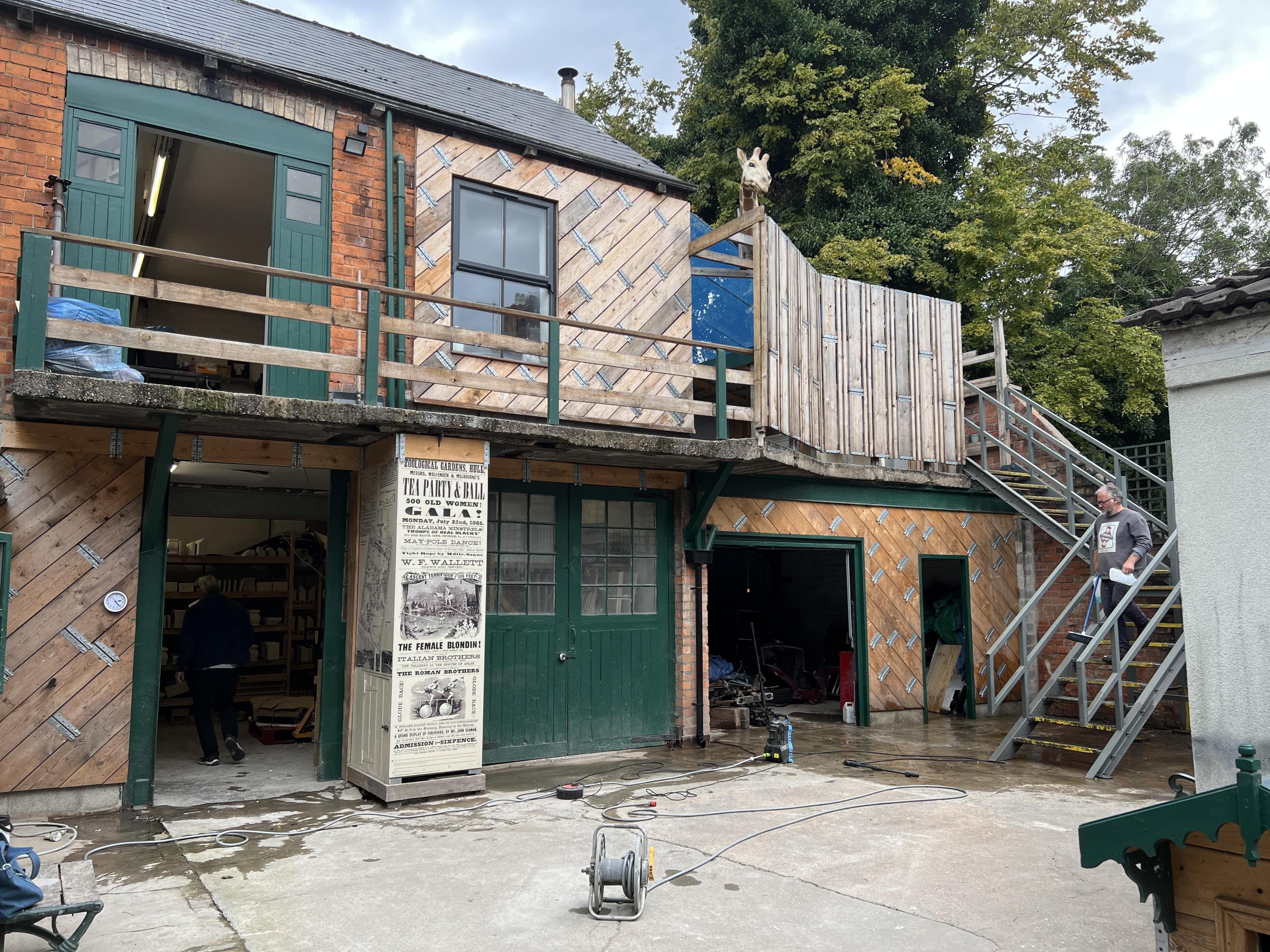 A two-storey building with a mix of wood cladding and brick. The ground floor has three green doors. A staircase leads to a balcony on the upper floor.
