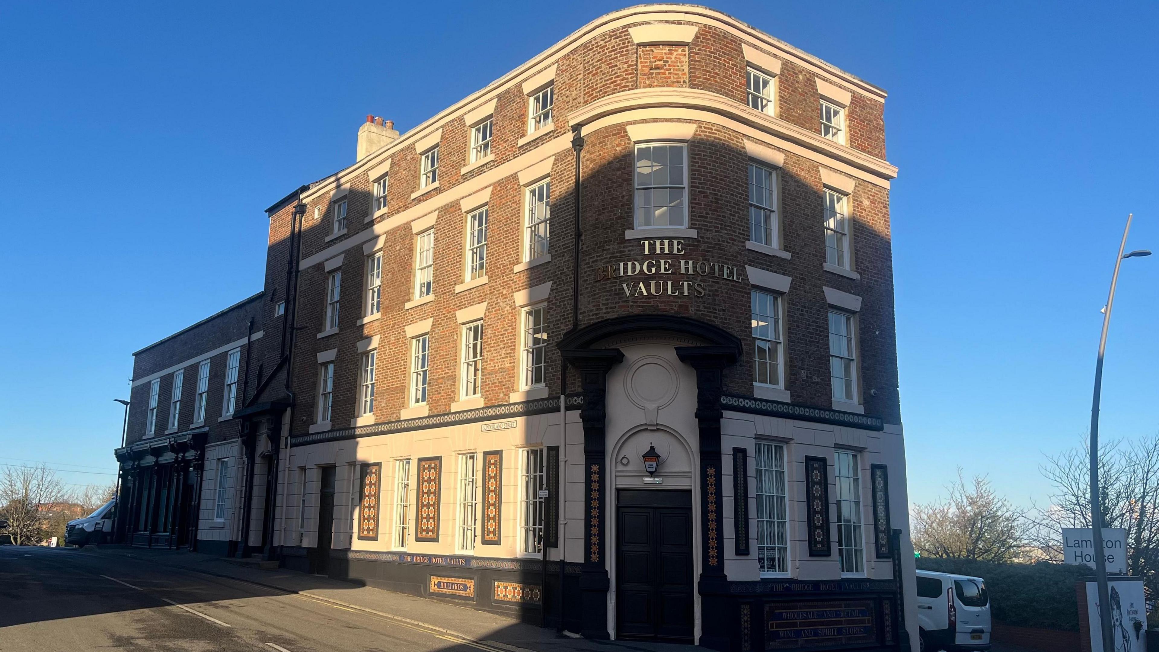 A grand four-storey building. It says 'The Bridge Hotel Vaults' in gold lettering on the front, and the ground floor has white brick with mosaic tiles. The rest of the building is made from red brick. There is an old fashioned lamp above the door.