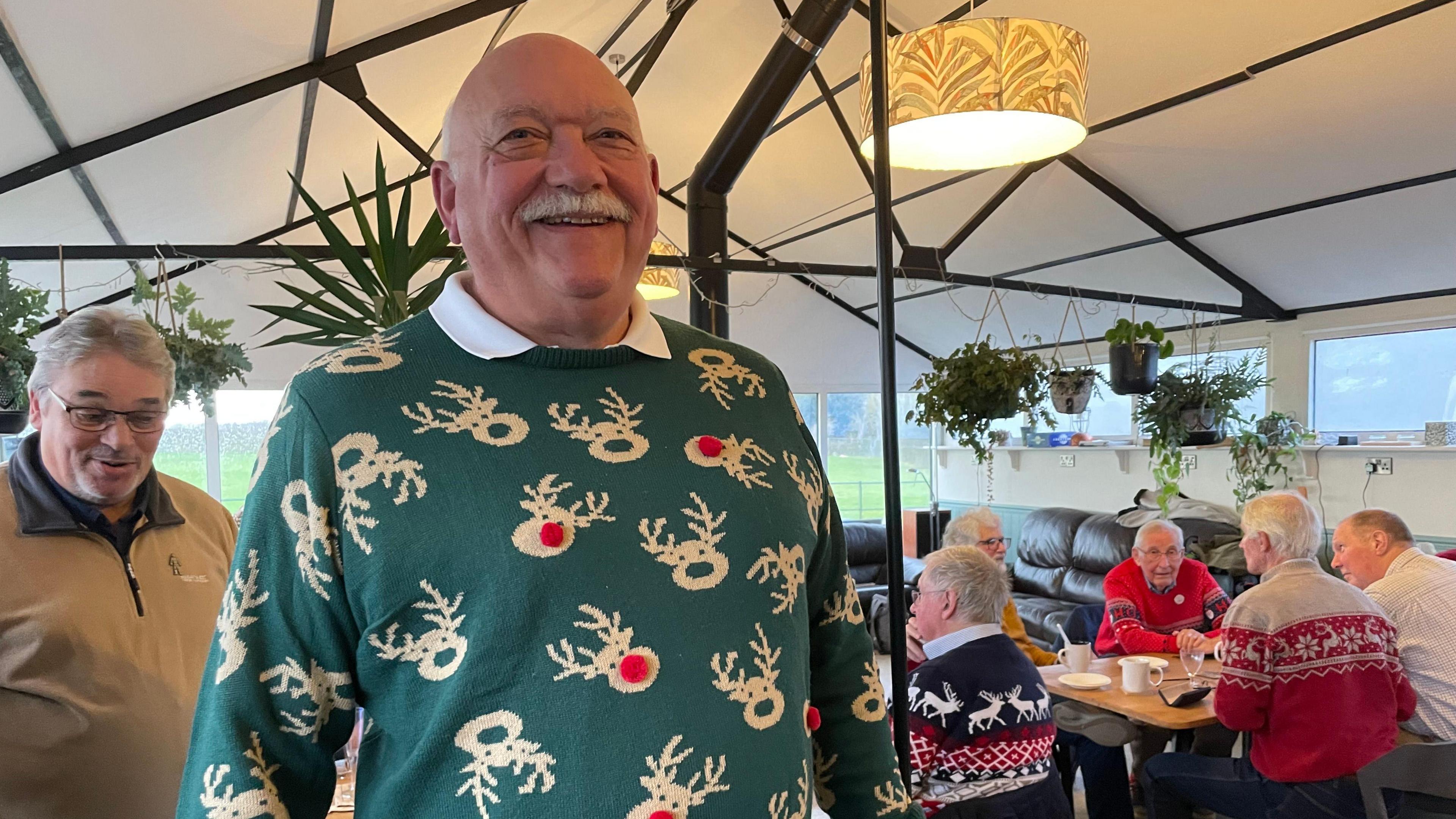 Graham is bald and has a grey moustache. He is smiling and is wearing a green jumper with brown reindeers on it. Some of the reindeers have red bobbles for noses. In the background, men with Christmas jumpers are sitting at a table.