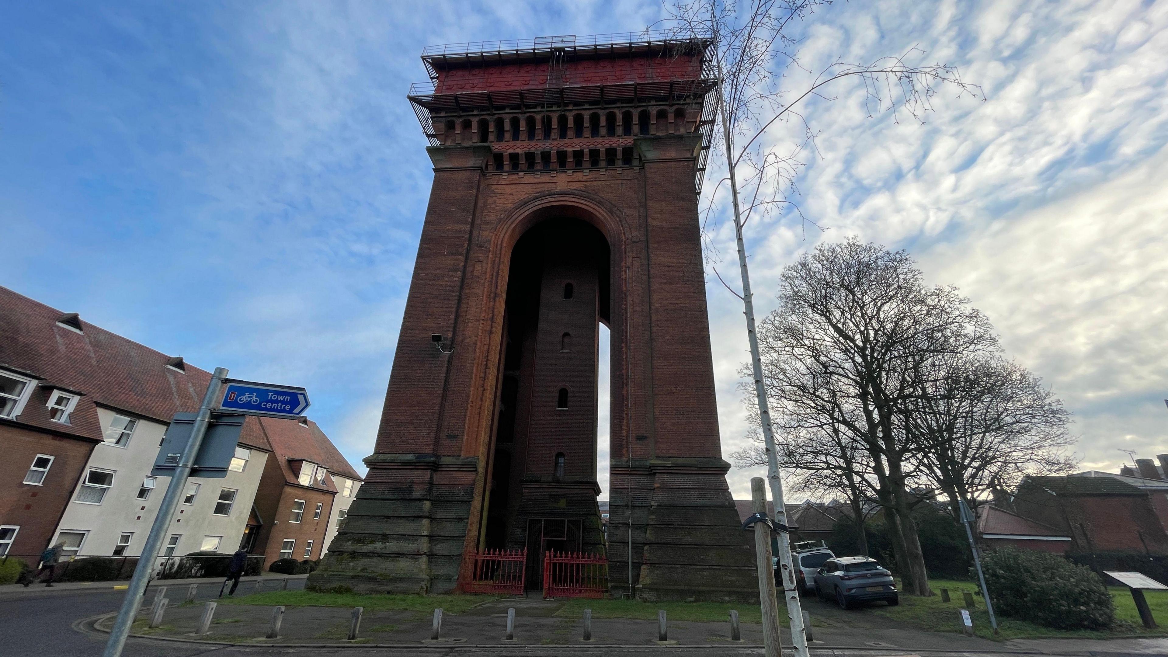 An imposing water tower built out of bricks looms over a grass area, some small bollards, some houses and cars. 