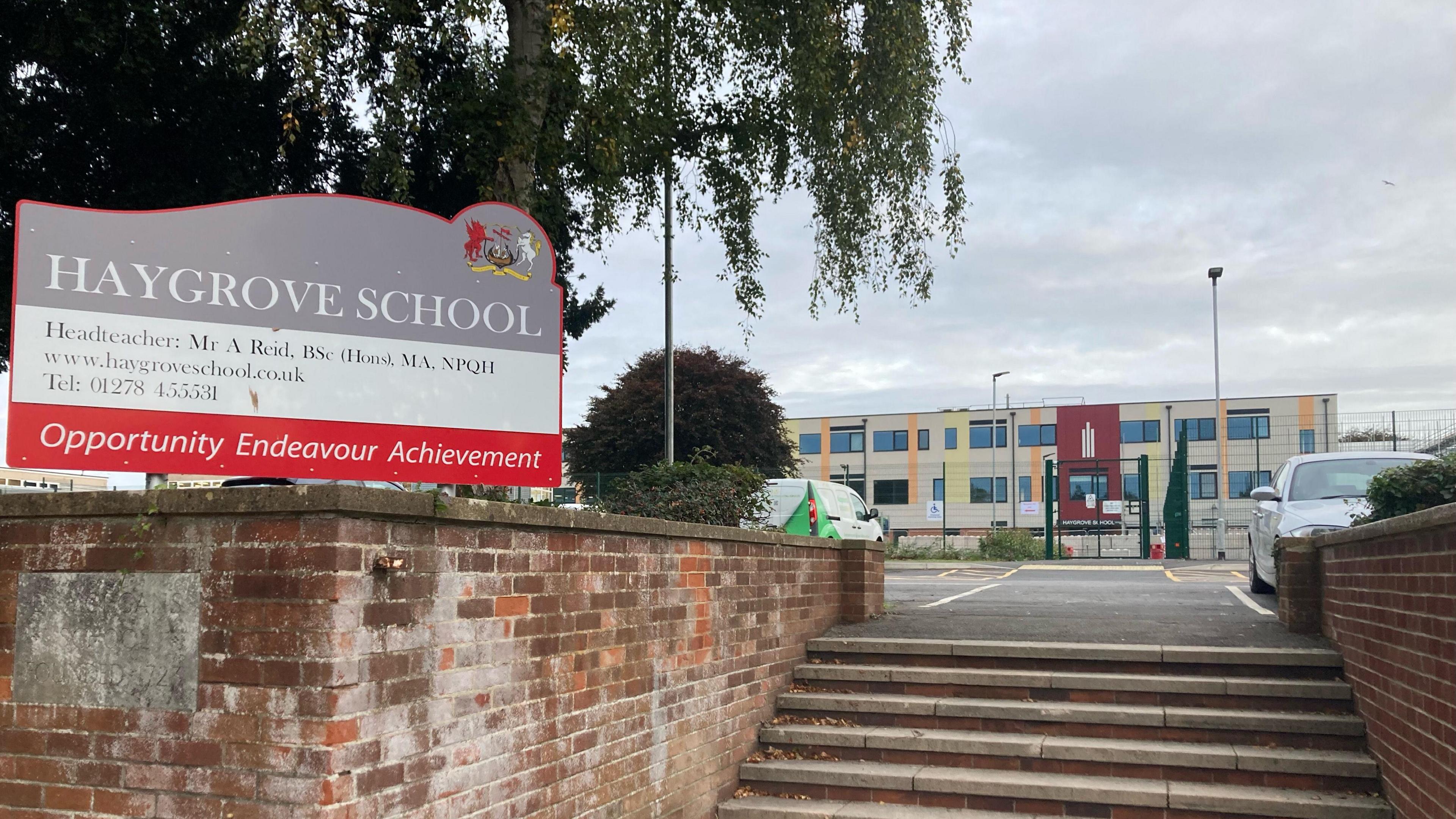 Haygrove School sign with a red brick wall. There's steps up towards a car park and then a building in the background.