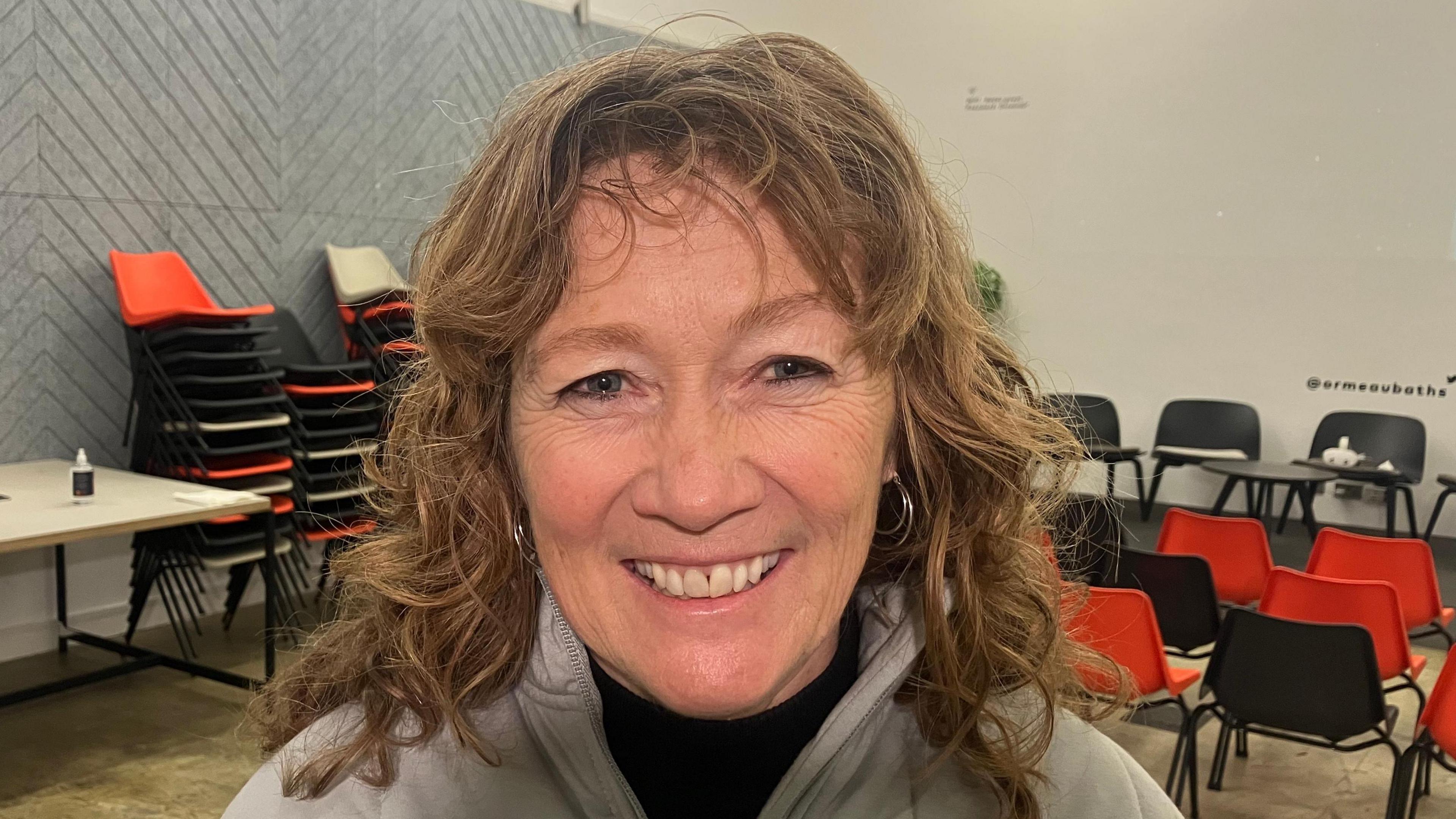 Professor Craig with shoulder-length brown curly hair. She is smiling at the camera as she wears a grey coat and black top. In the background rows of red and black plastic chairs can be seen.