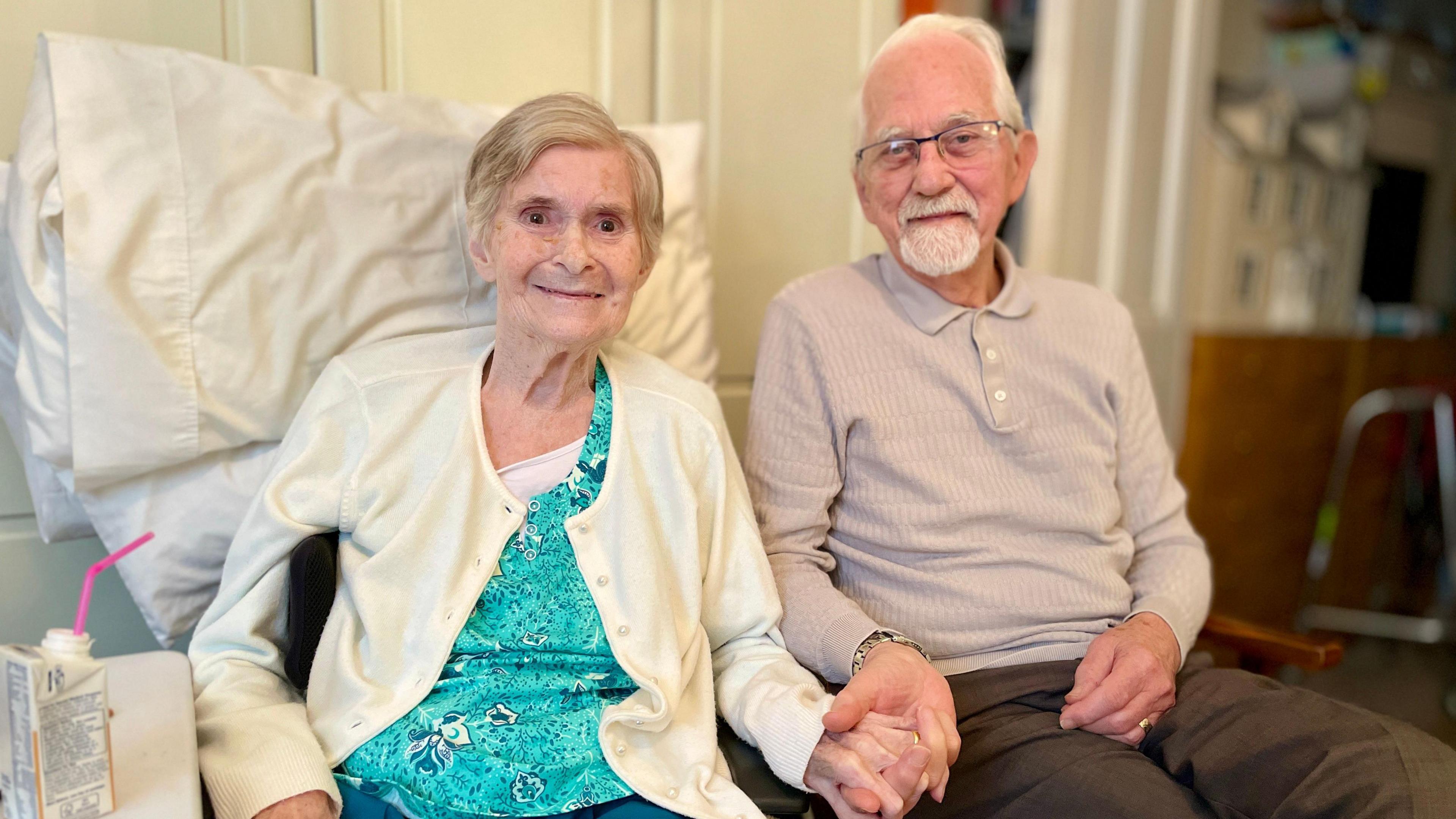 A man with white hair and a white beard, dressed in a grey/brown jumper and brown trousers, sits holding hands with a woman with grey hair and a white cardigan who is propped up with a pillow.