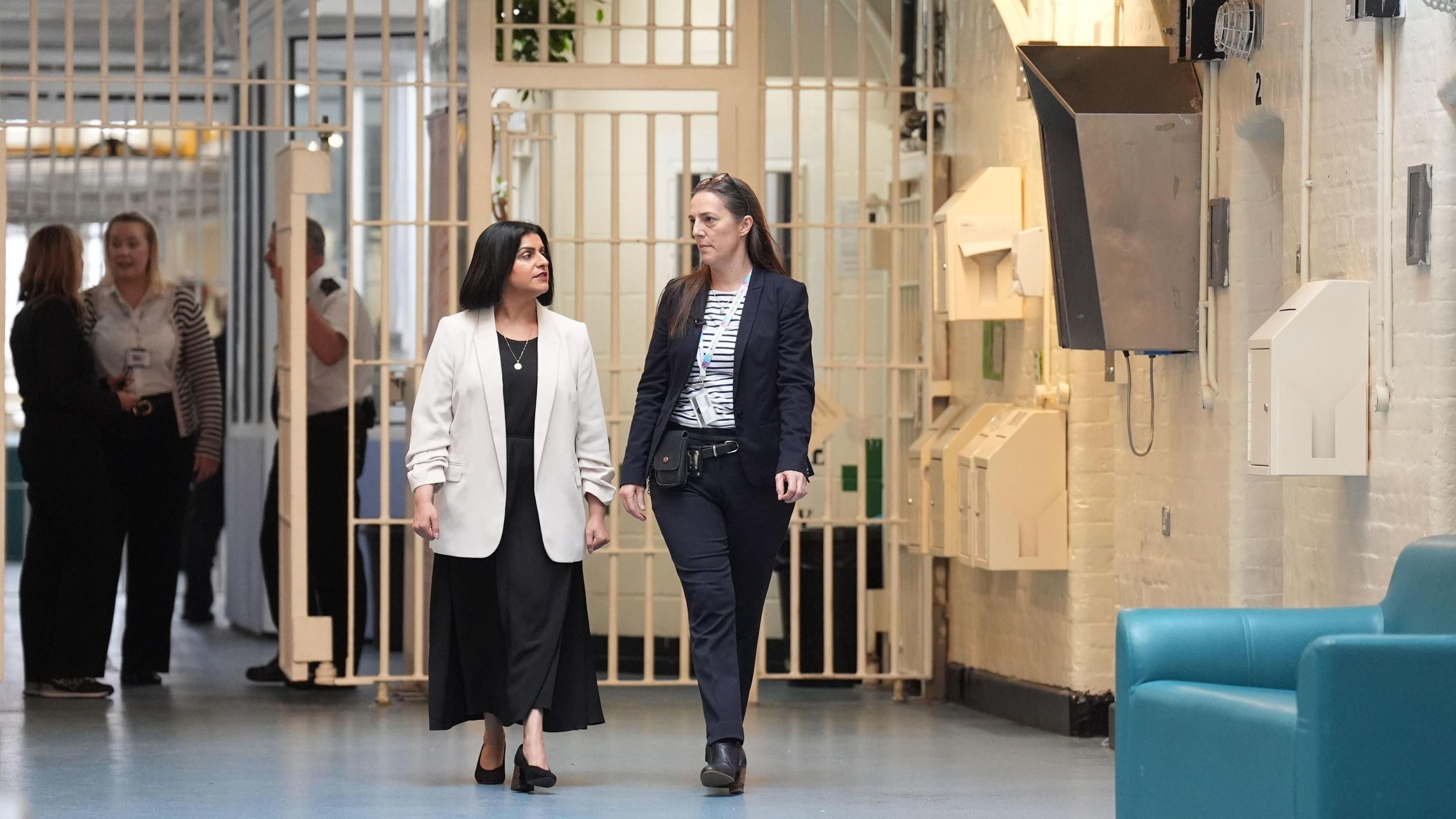 Justice Secretary Shabana Mahmood, wearing a black dress and white blazer, is walking and talking with Sarah Bott. who is dressed in black trousers and blazer with a striped top underneath. Staff can be seen chatting in the background behind a wall of white prison bars.