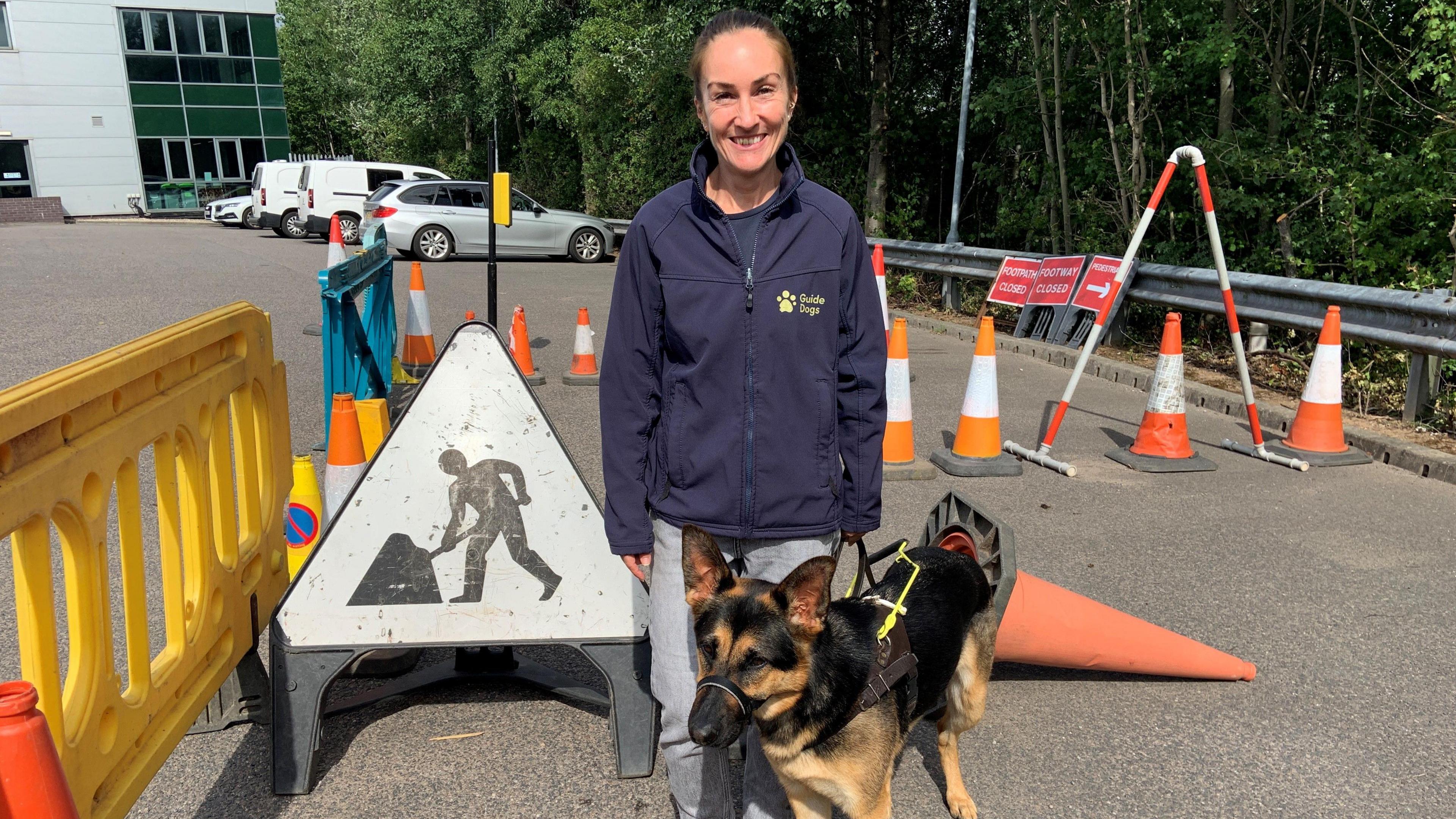 A woman is wearing a blue fleece with a yellow logo saying "guide dogs" She is standing in a mock street with fake roadworks and has an Alsatian guide dog on a harness by her side