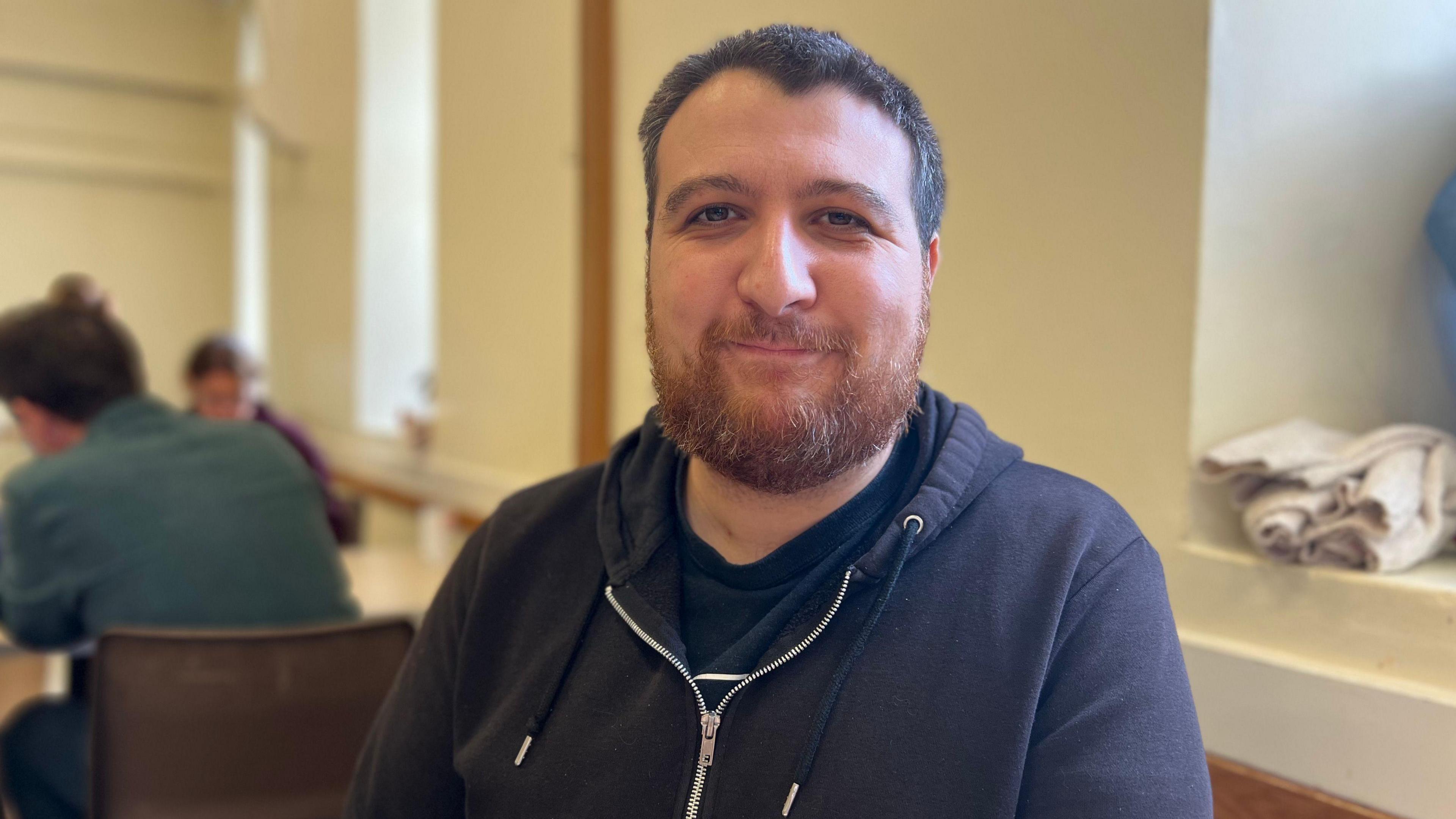 Tamer pictured with short brown hair and a beard. He is wearing a navy zip up hoodie.