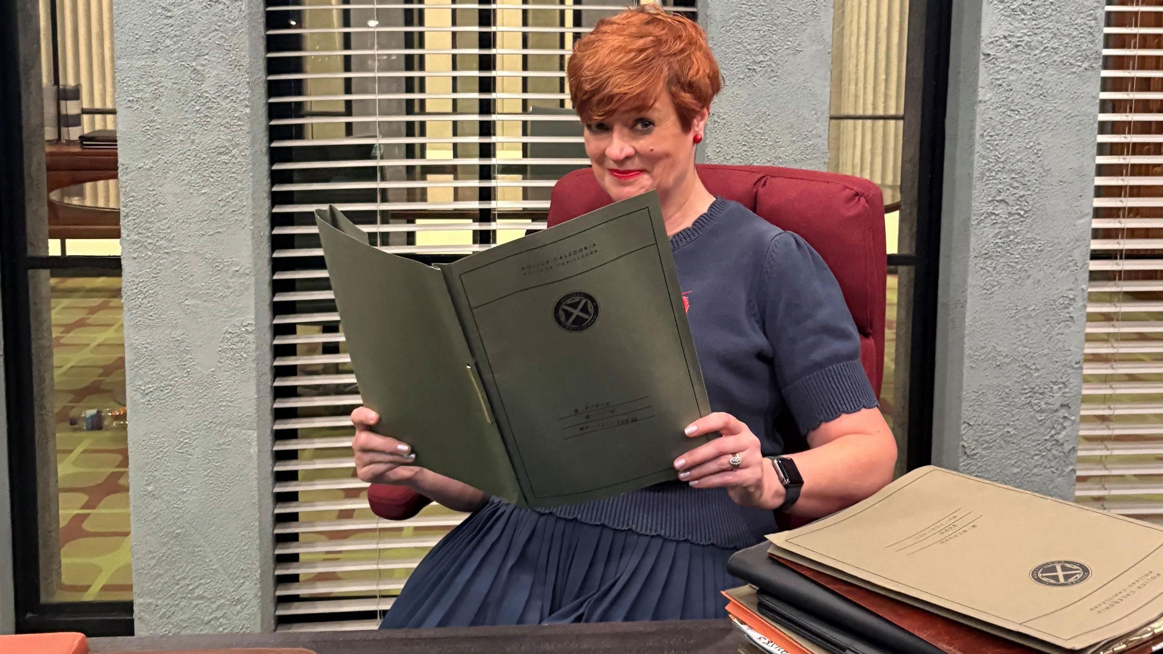 Pauline McLean sitting behind a police desk holding a crime report. There are blinds on windows behind her with a room behind