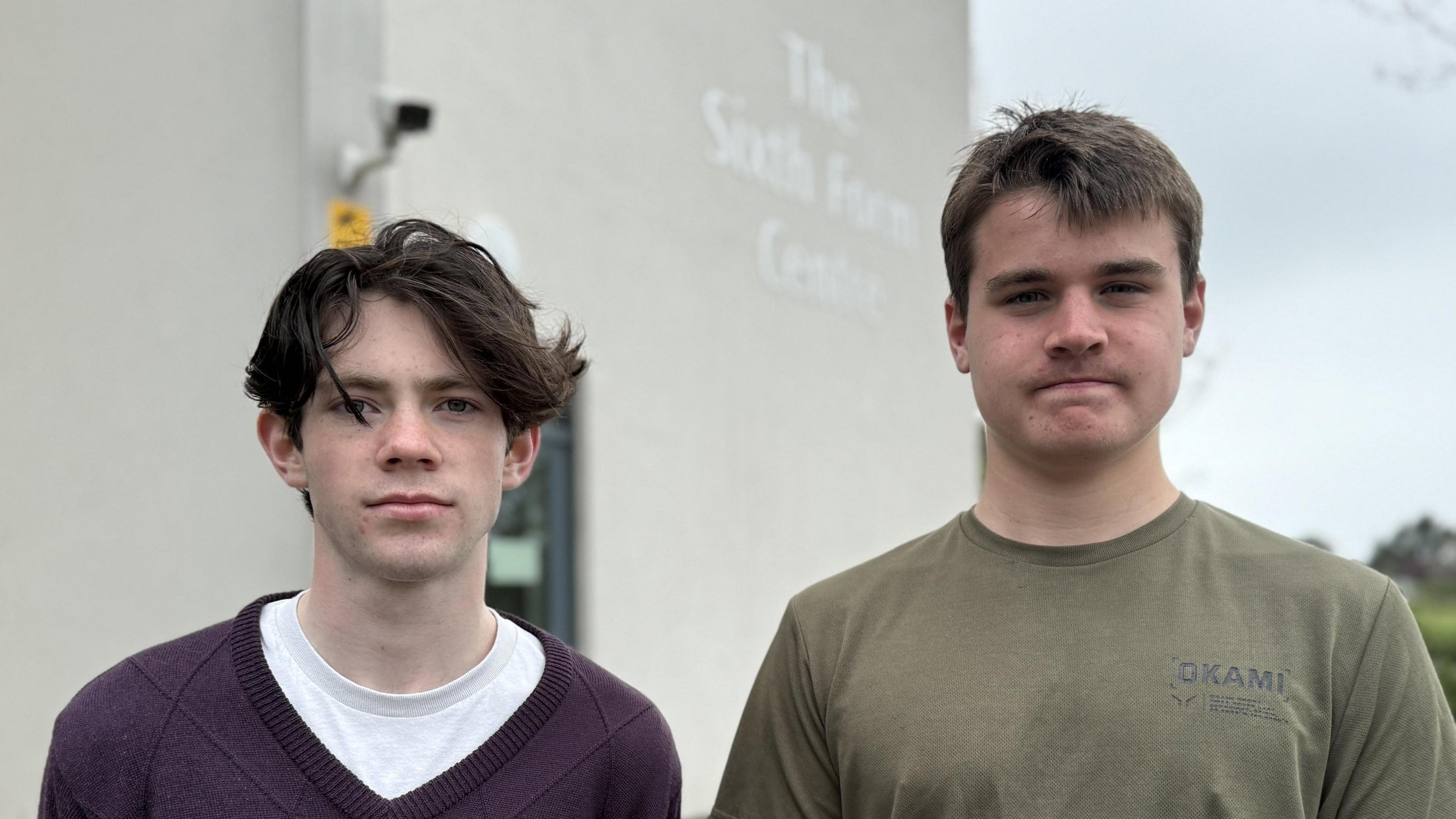 Two teenage boys looking at the camera, with the Guernsey Sixth Form Centre in the background. The boy on the left, Ben, is a white boy with medium length dark brown hair with a parting down the middle. He is wearing a dark purple V-neck sweater with a white t-shirt underneath. The boy on the right, Shea, is a white boy with shorter, mousey brown hair. He is wearing a khaki green t-shirt.