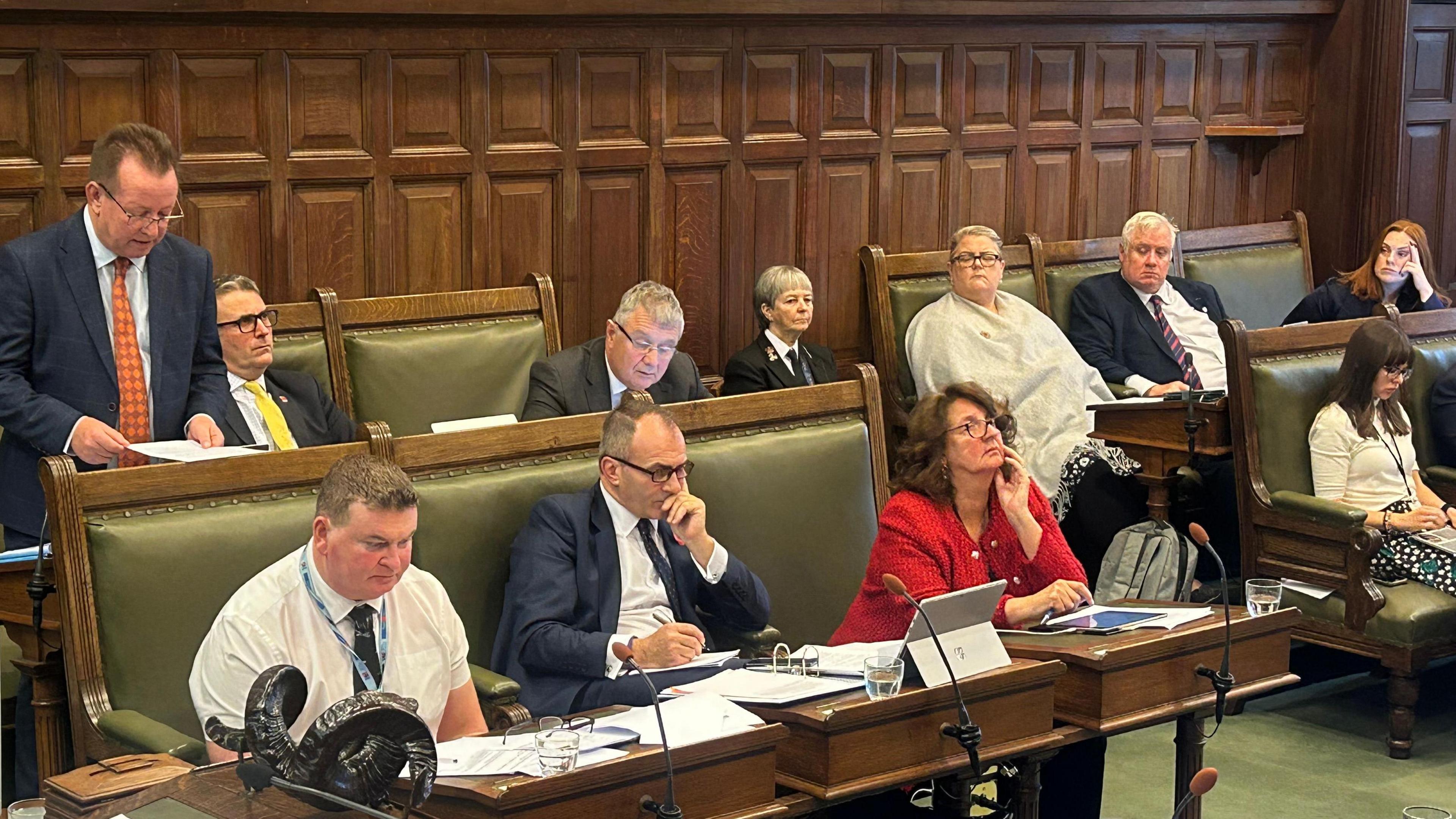 In the House of Keys, an old wooden panelled room with wooden benches, members sit along the benches. Tim Glover stands up as he moves the motion and Alfred Cannan sits on the row in front looking down.