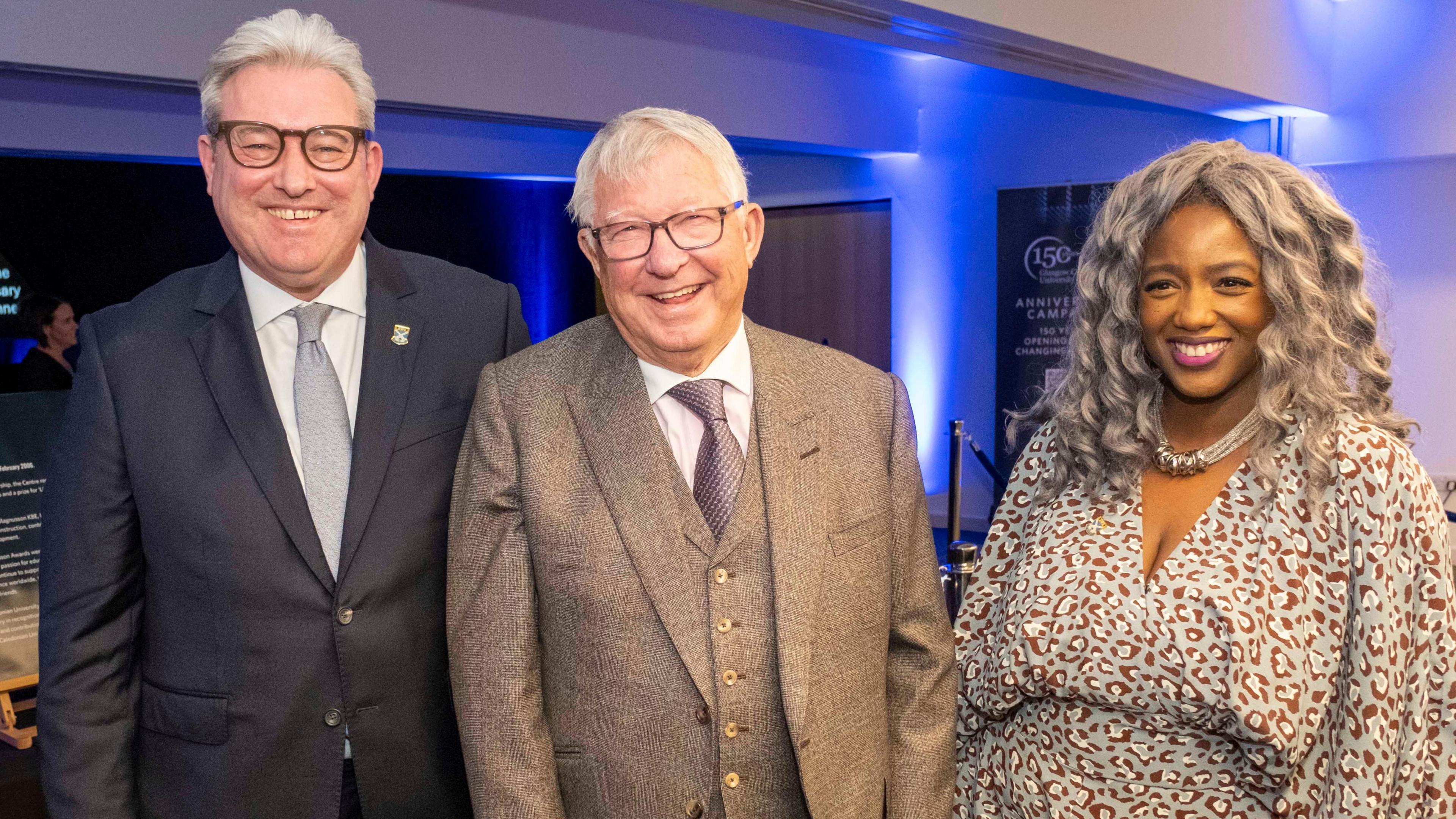 Dr Murray Rowden, Sir Alex Ferguson and chancellor Dr Anne-Marie Imafidon pictured smiling while standing alongside. Both men have grey hair, dark-rimmed glasses and are wearing suits, white shirts and ties. Dr Imafidon has long grey curly hair and is wearing a blue, brown and white patterned dress.
