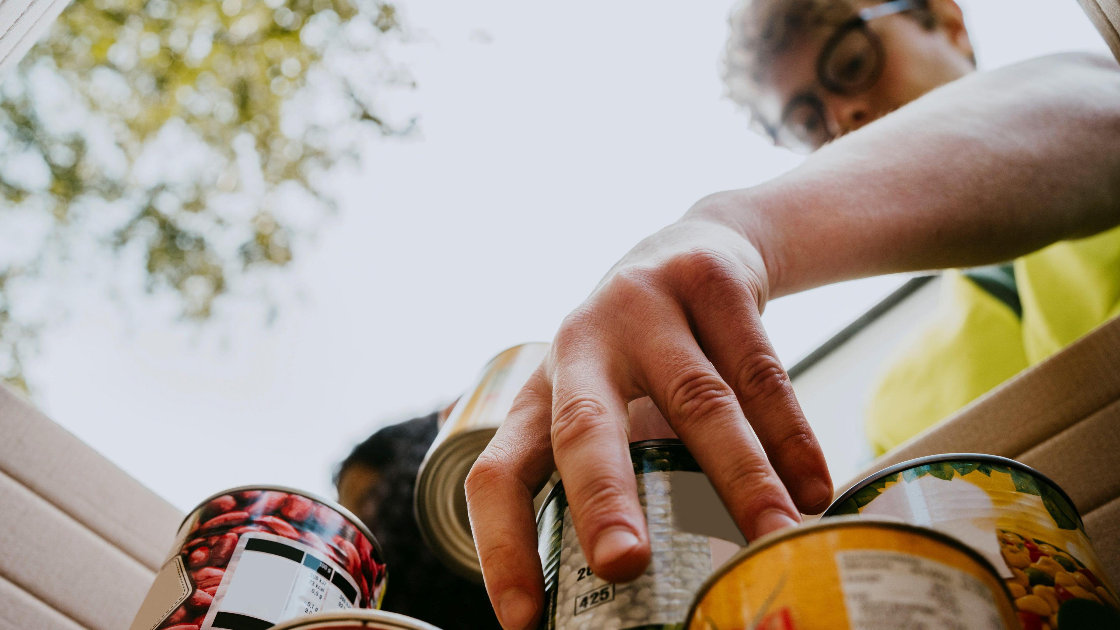 A man wearing glasses is looking down at a box filled with canned food. He is wearing a bright yellow jacket and holding one of the cans. Trees are above him.