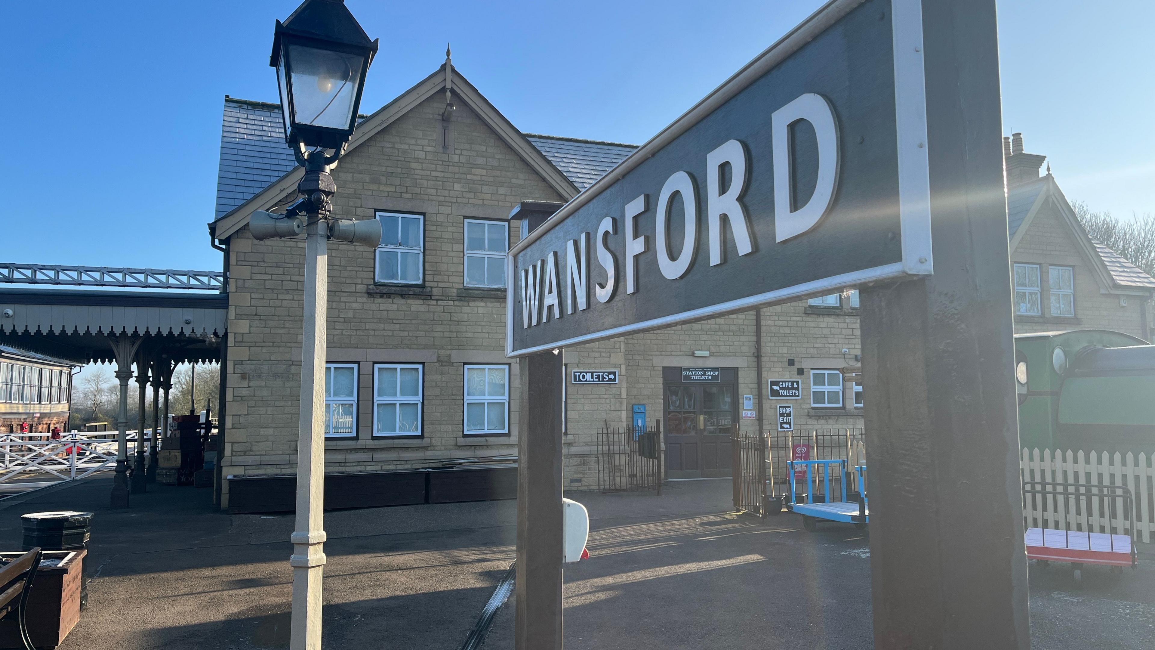 A sign on the NVR platform reads Wansford. Behind it is the station building and the train tracks are situated to the left. 