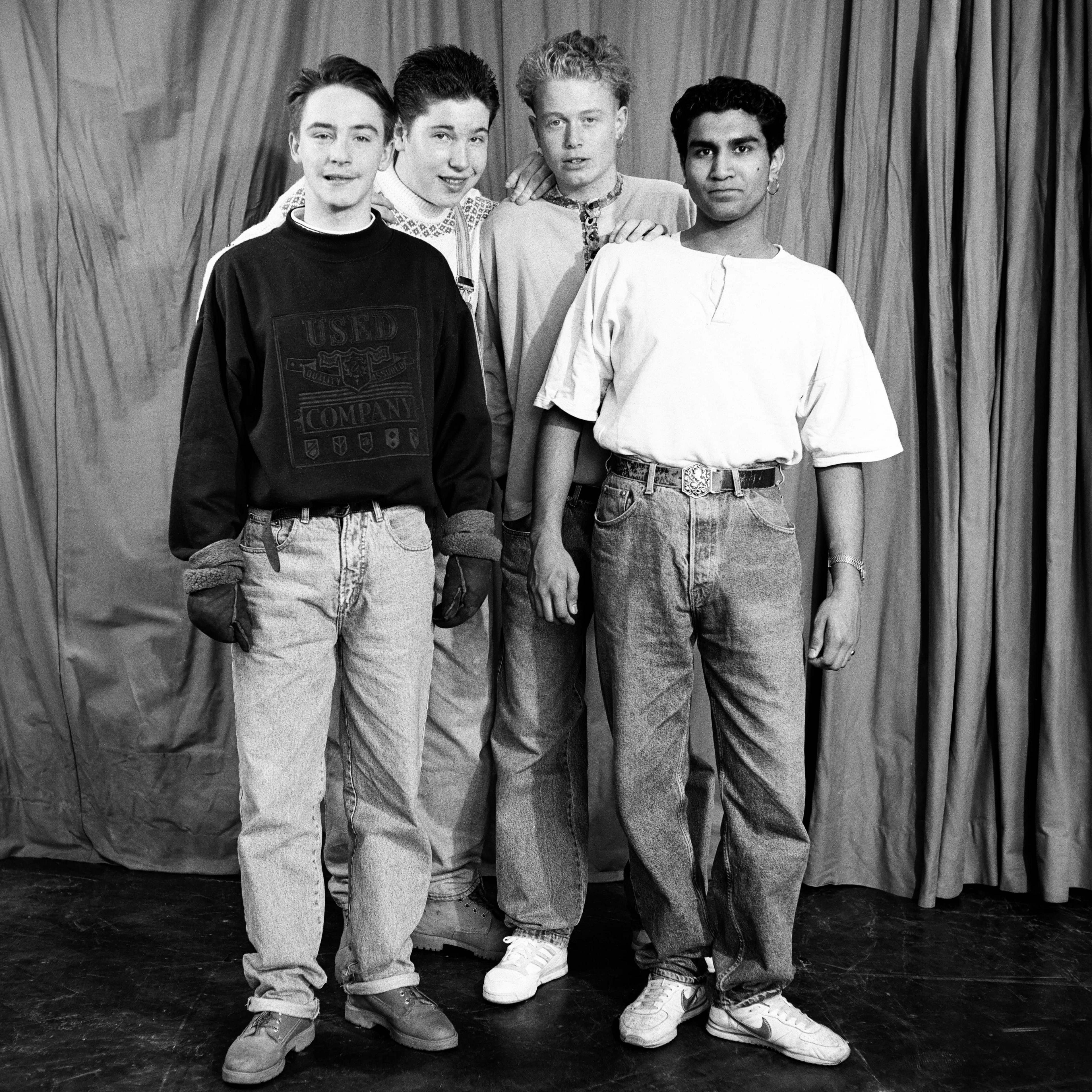 Boys from Tulse Hill School, 1990. Four young men stand in front of a curtain. They wear wear jeans and casual tops. Black and white photo.