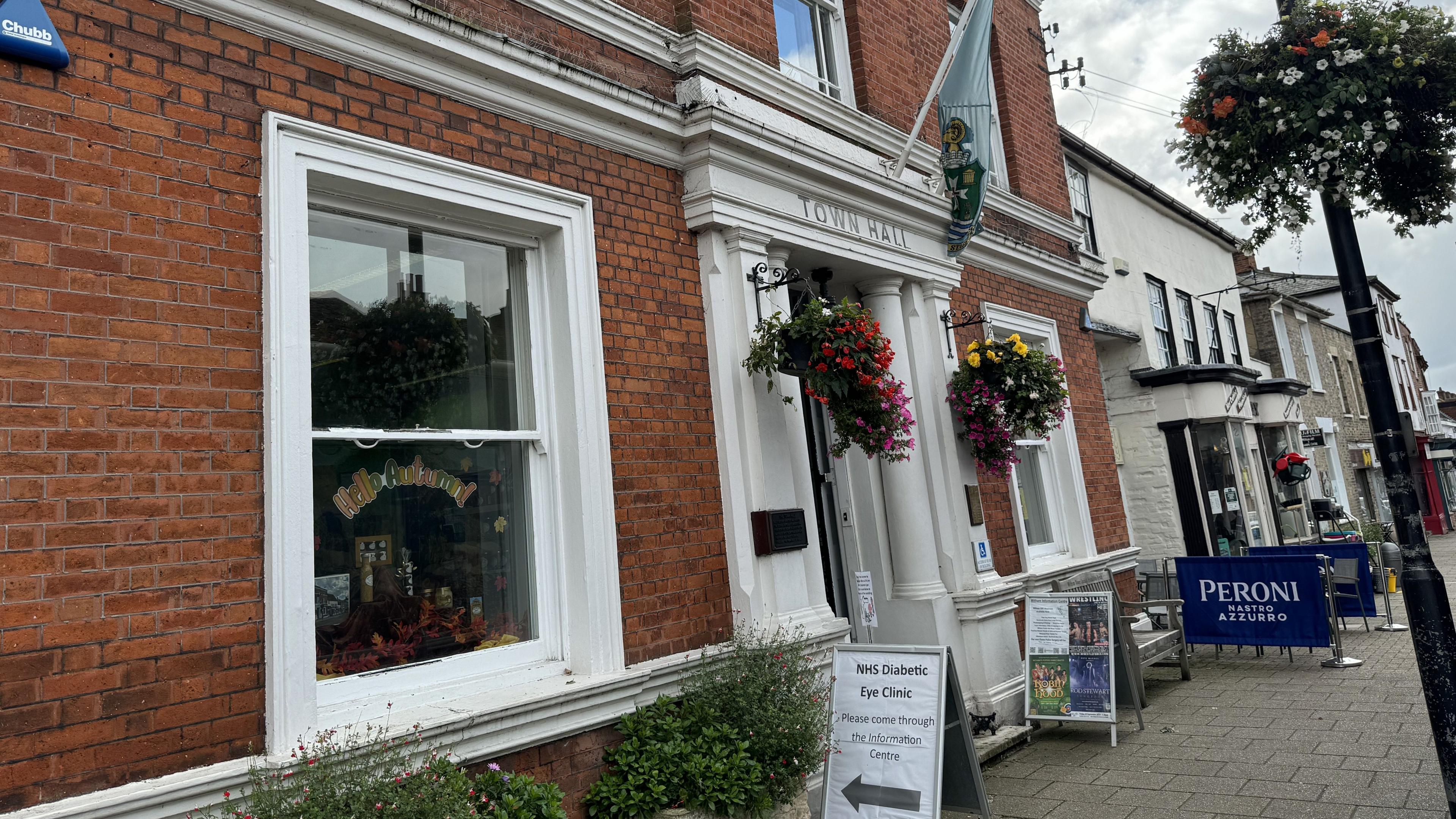 Outside Witham Town Hall. The entrance is white with the words "town hall" in black above the door. There are hanging baskets on either side of the door and a flag above it.