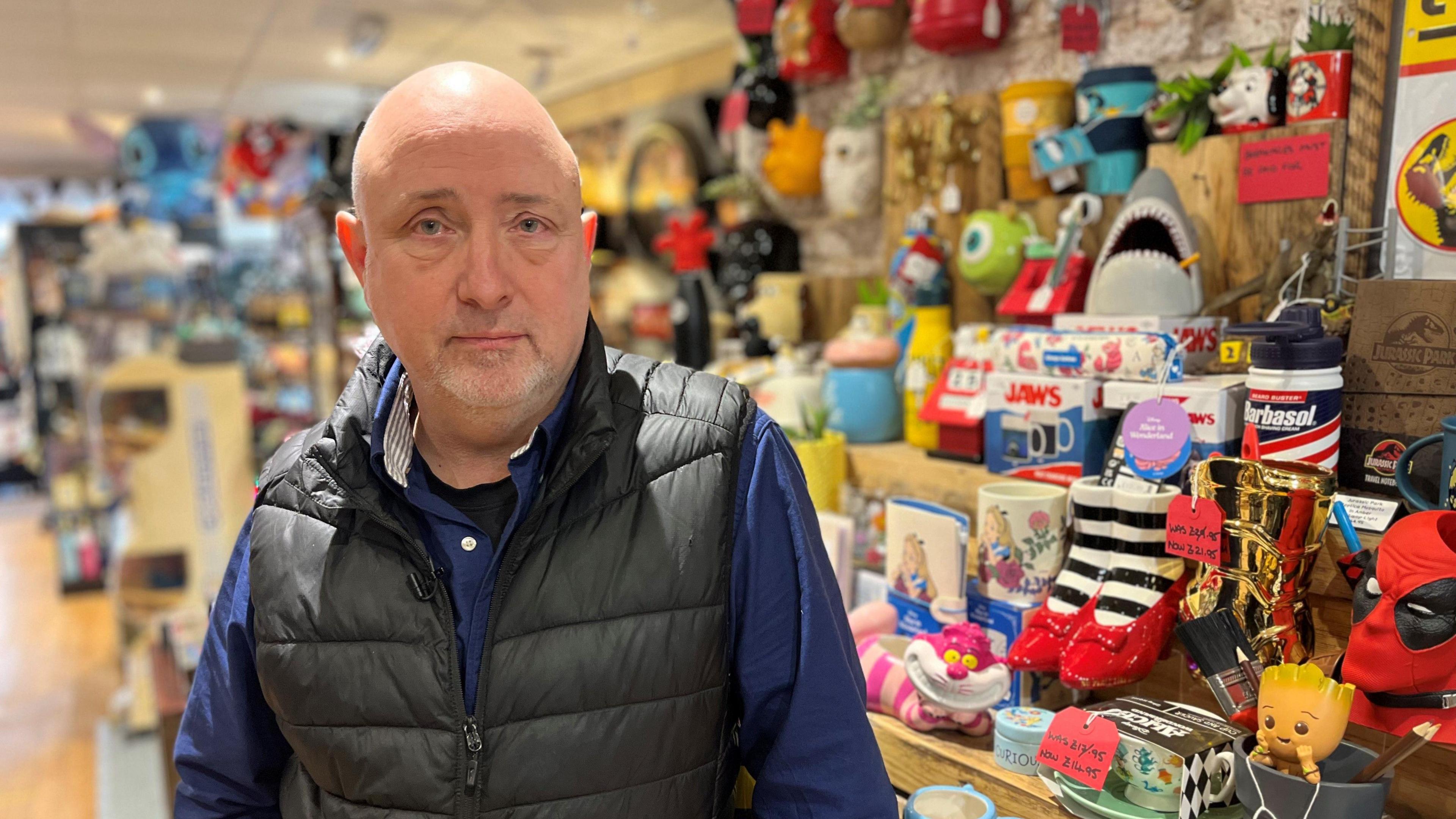 Martin Gaunt, aged 62, wearing a blue shirt and black padded gillet, with colourful stock on shelves in the background.