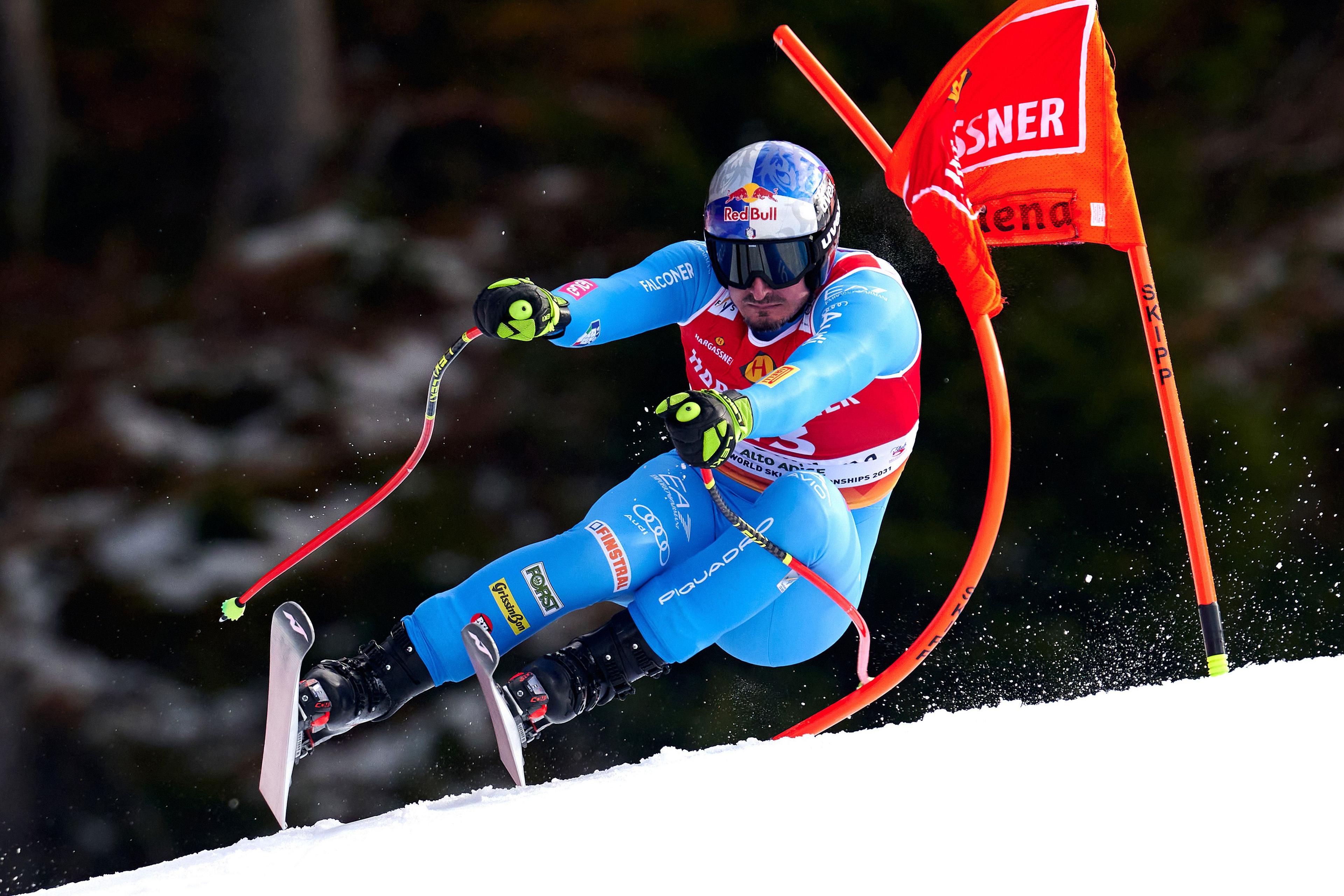 A skier in a blue racing suit and helmet speeds downhill during an alpine skiing event, leaning into a turn while passing an orange gate flag. The skier’s poles are angled outward, and snow sprays up from the skis as they carve through the course. A forested background is softly out of focus behind the action.