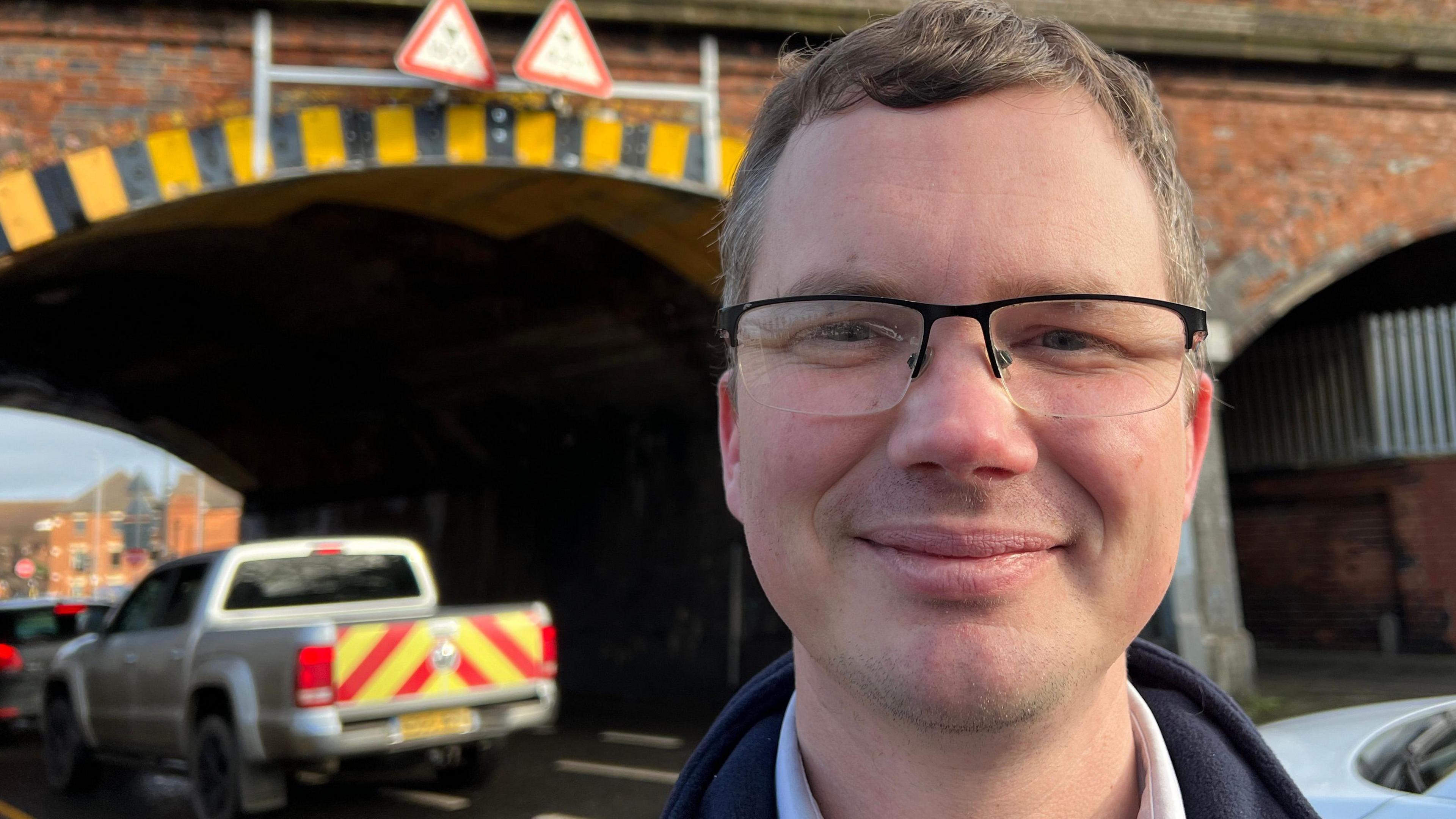 Michael Clegg is standing near an arched brick-work bridge as a van passes beneath it. He has light brown hair, is wearing glasses and is smiling with his lips closed.