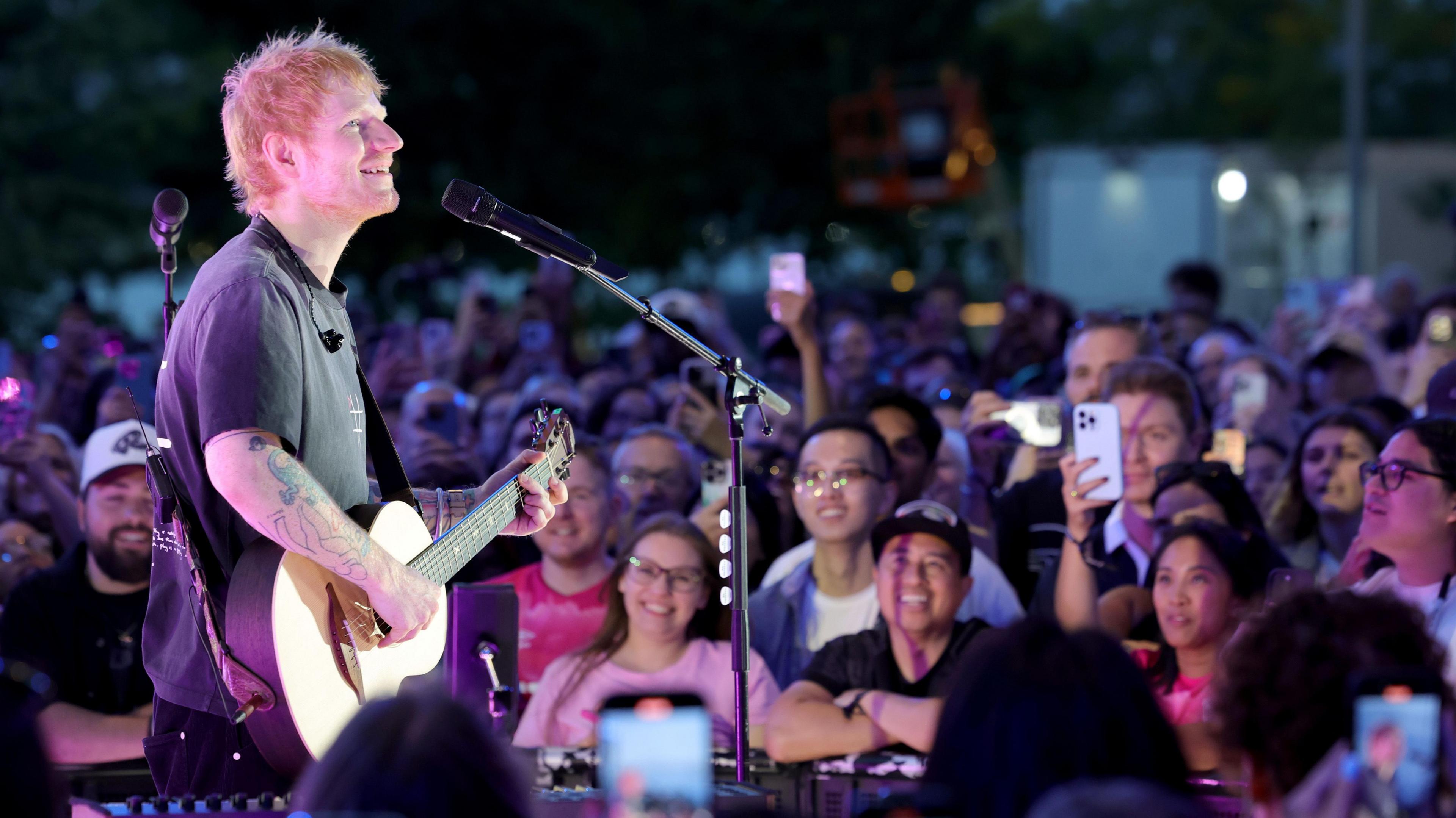 Ed Sheeran plays guitar and sings in front of a crowd, smiling and filming on their phones.