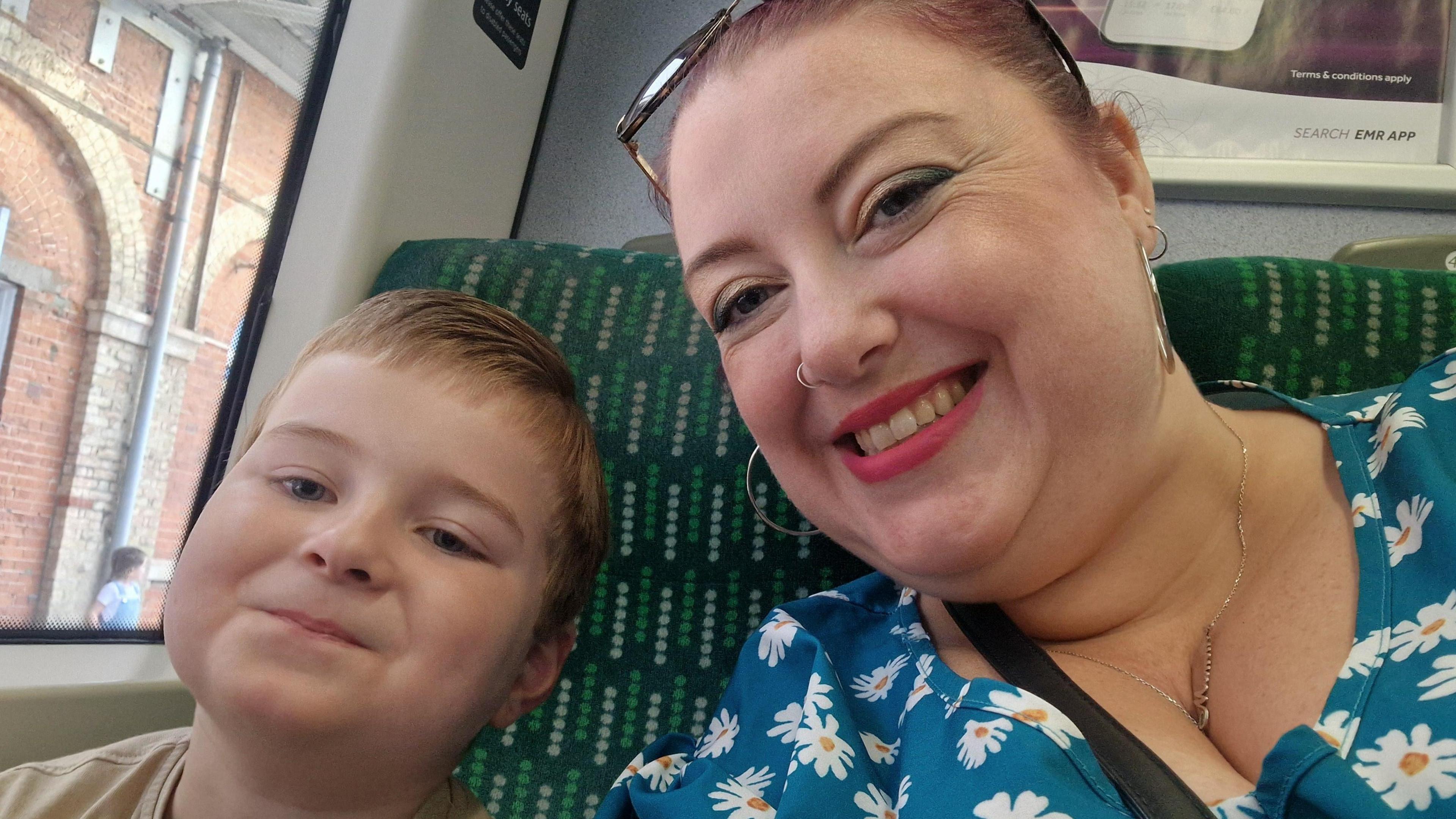 A selfie picture of a mum and her seven year old son inside a bus. The mum on the right is smiling and wearing a blue dress with daisies on, and wearing sunglasses on her head. Her son is smiling and wearing a grey-green top. 
