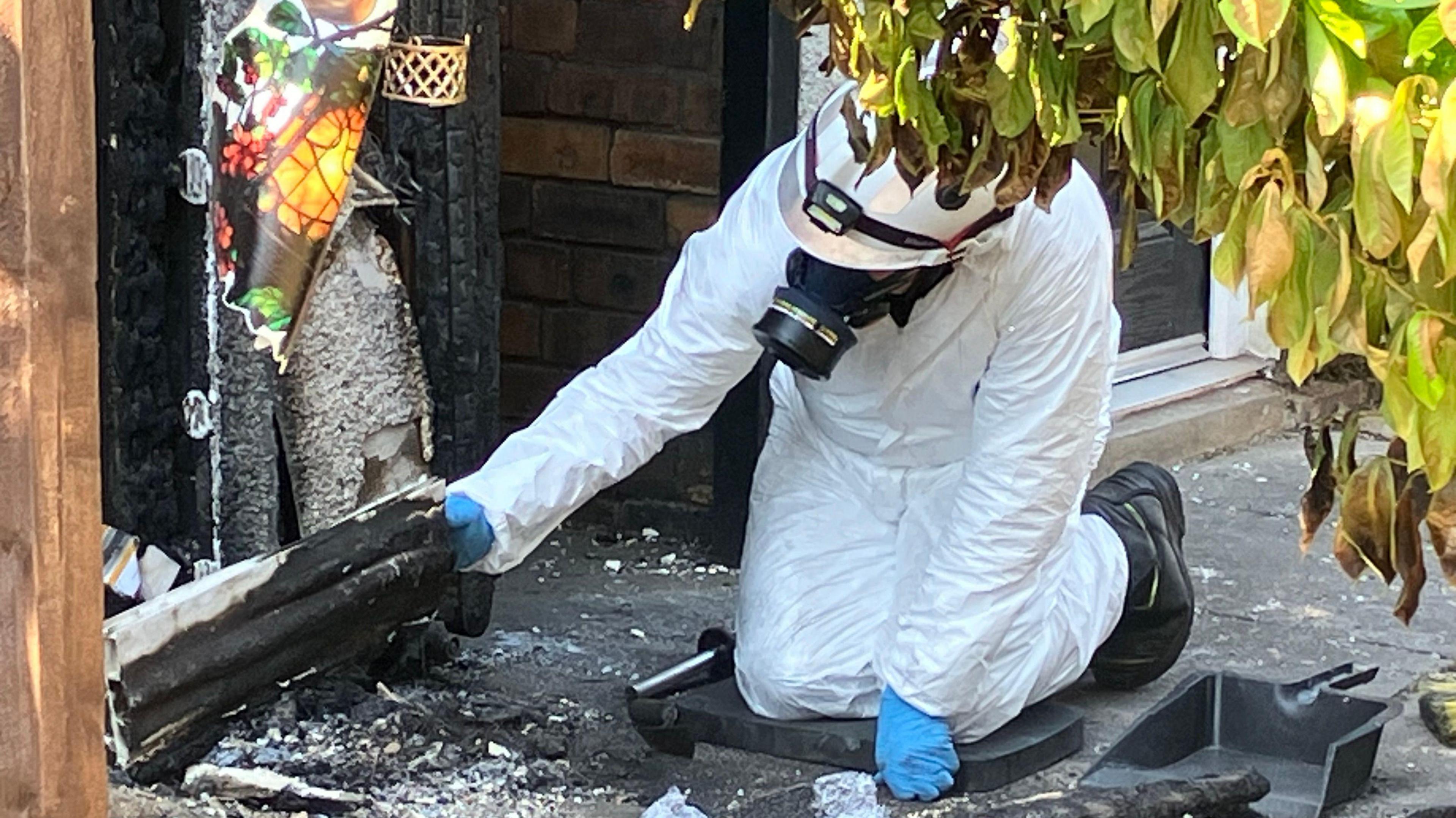 A person in a white forensic suit is kneeling down in the wreckage of a house fire and is lifting up a white panel that has also been damaged.