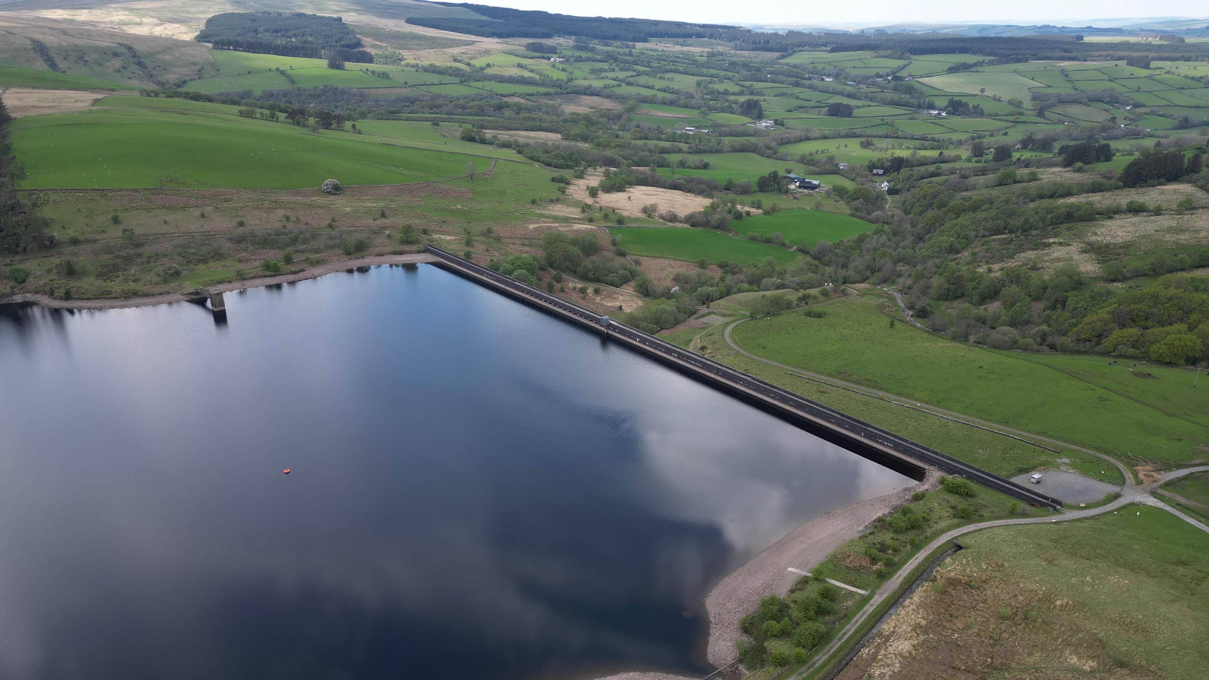Drone image of Crai Reservoir. A large body of water can be seen in the foreground, surrounded by hills, fields and trees