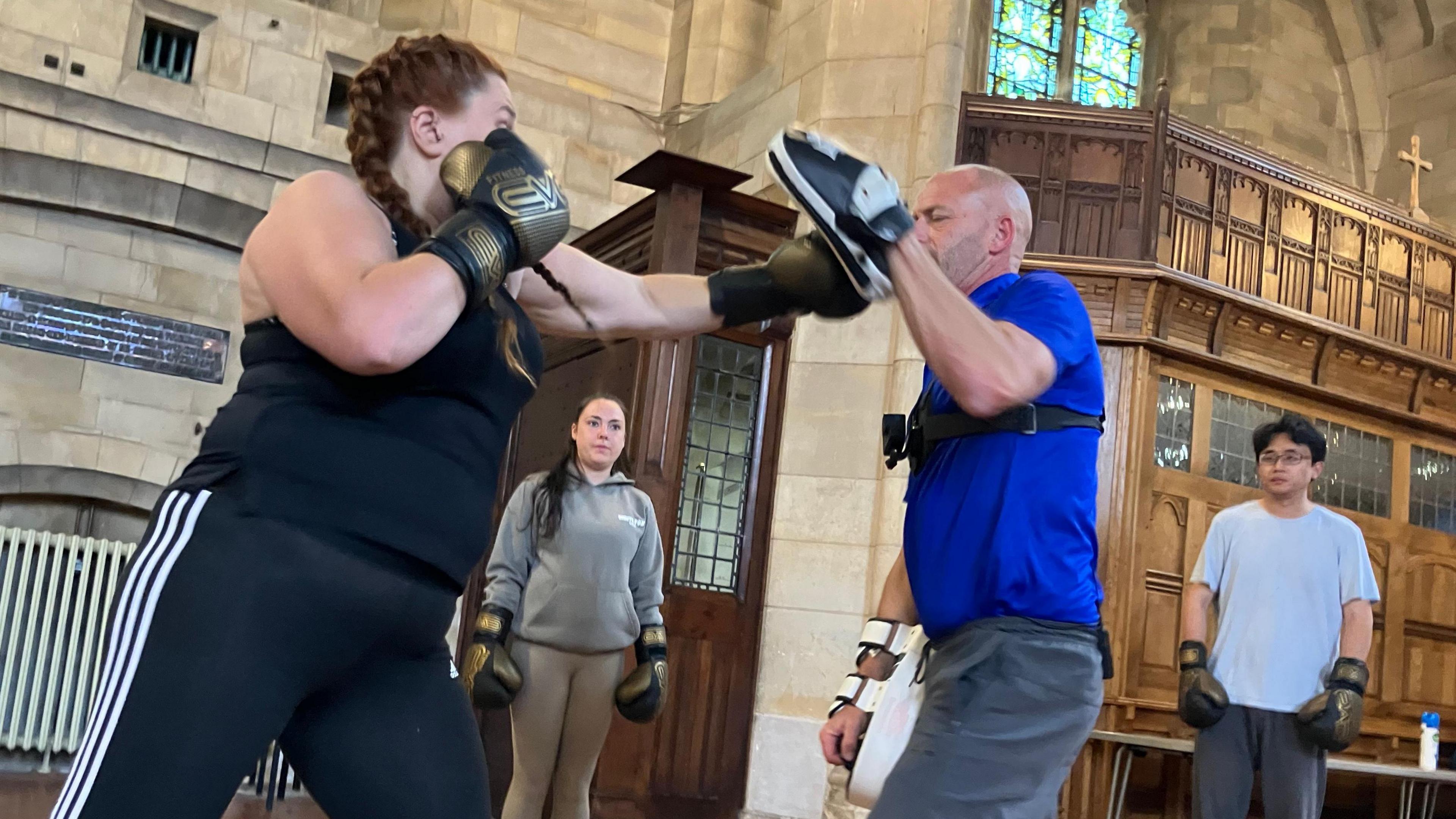 Ben Cookson takes a punch from a woman in black exercise clothes while wearing a pad on his left hand during a boxing class in a church. Two other people wearing boxing gloves are watching the action.