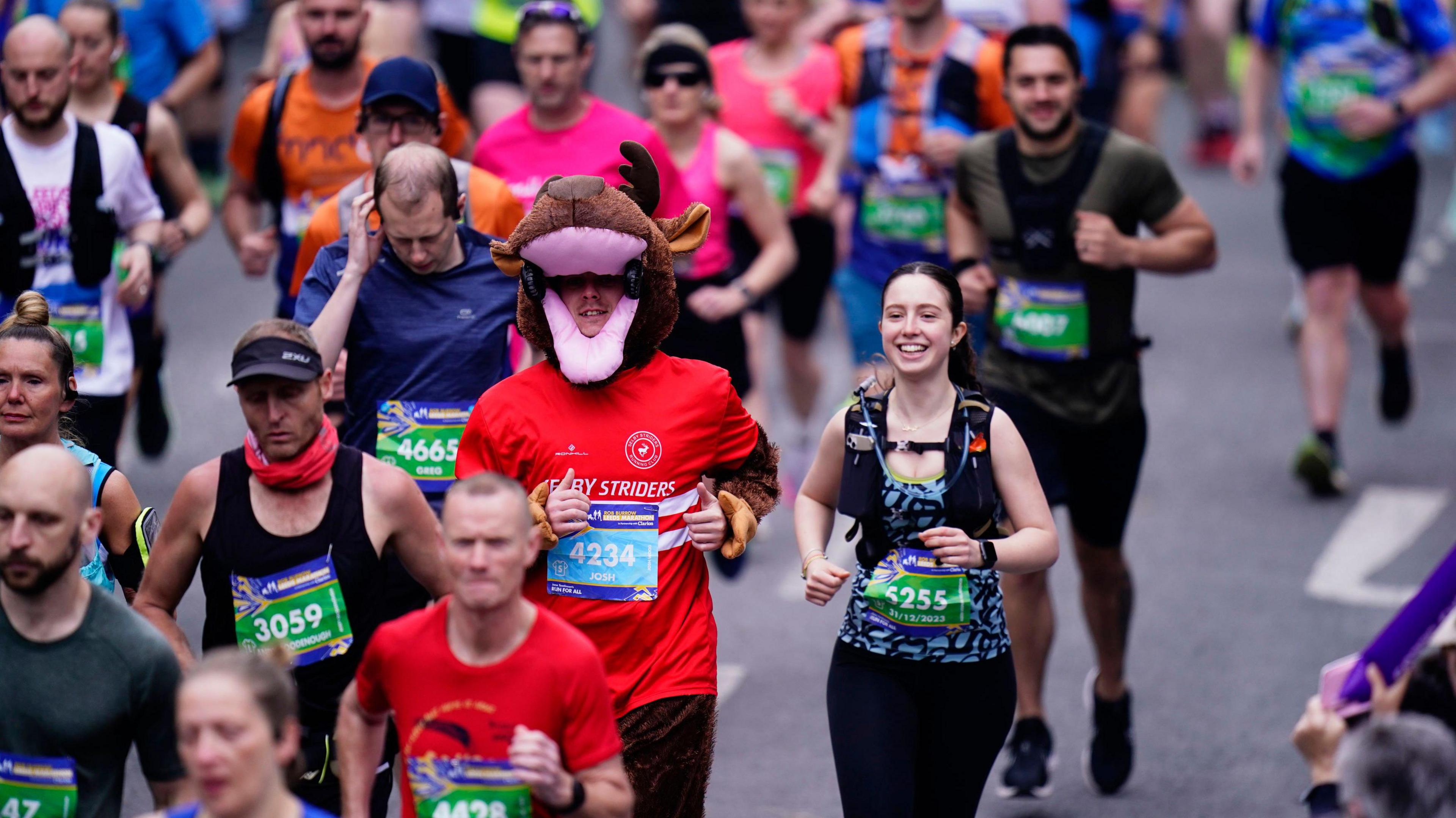 A crowd of people running through Leeds. One woman is in focus - she wears black and blue athletic gear and is smiling. A man in an orange top and animal mask runs next to her. Further runners are blurred wearing an array of athletic gear and their marathon numbers.