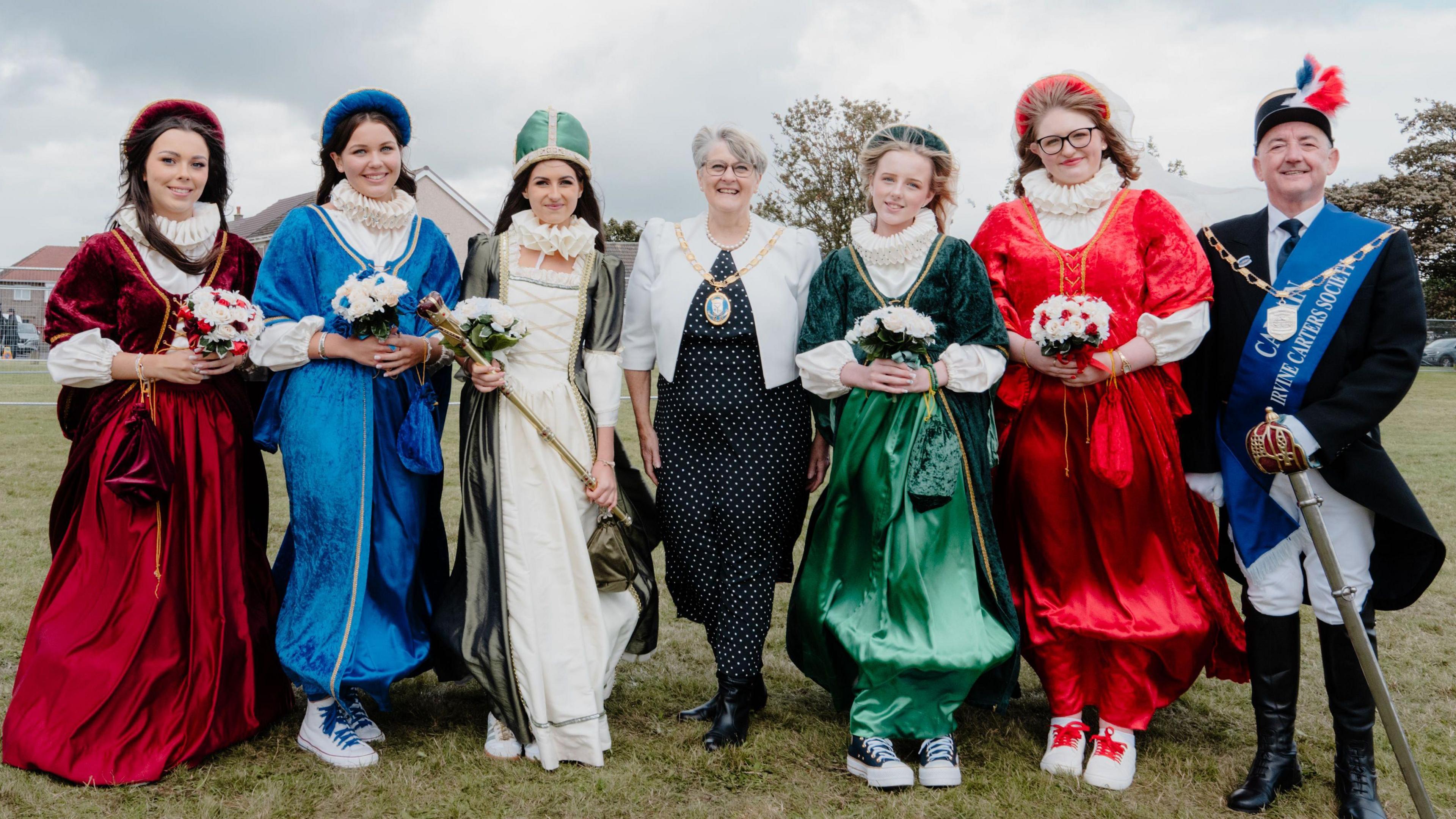 A group of seven people stand in a field and smile for the camera - five of the group are teenage girls dressed in medieval style dresses, and one has a crown on. The other two people are a man and woman, presumably local dignitaries - the man has a blue sash on as well.