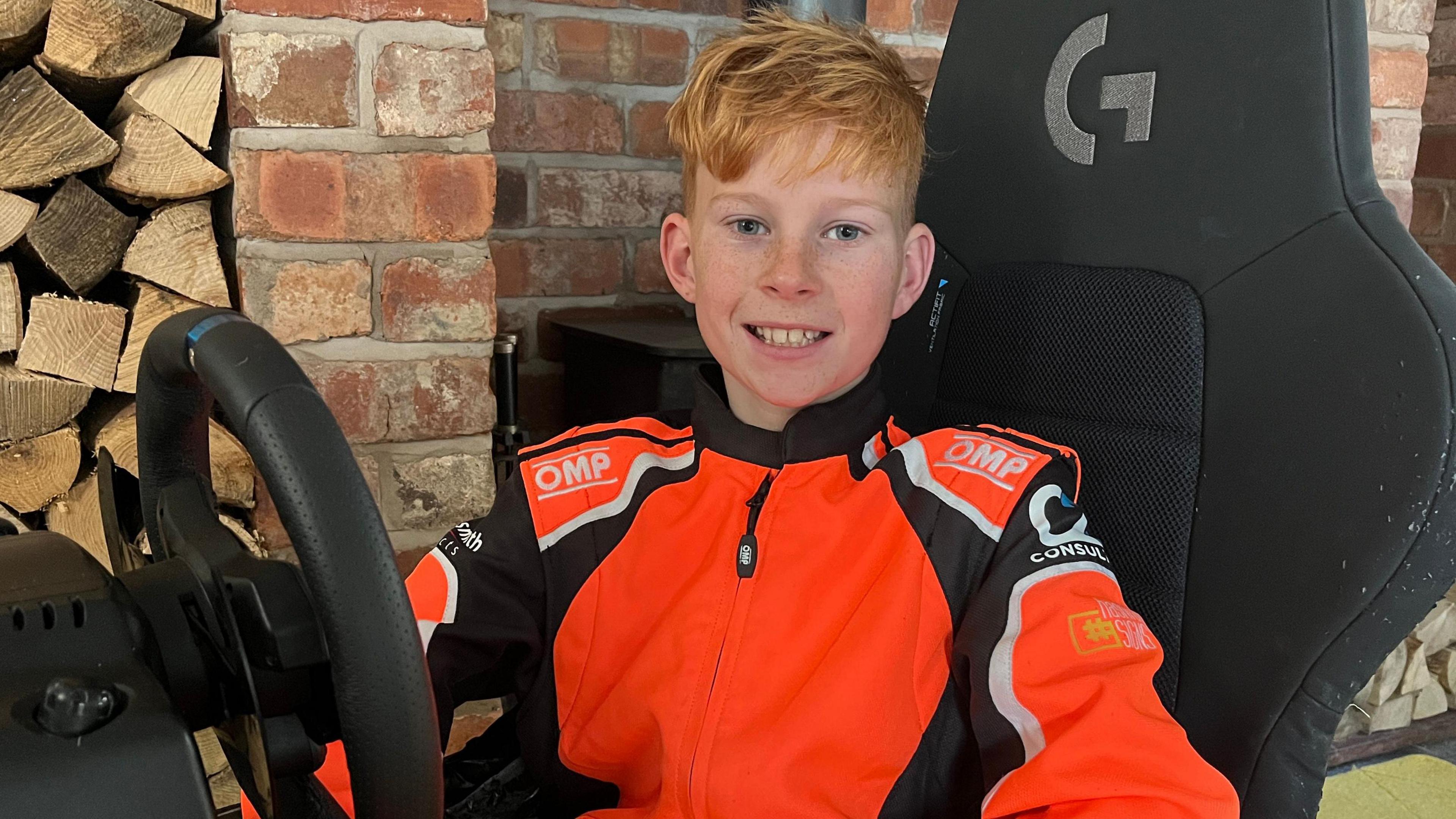 Zachary wearing an orange karting jacket. He has ginger hair and is sitting in a kart with a black seat and steering wheel