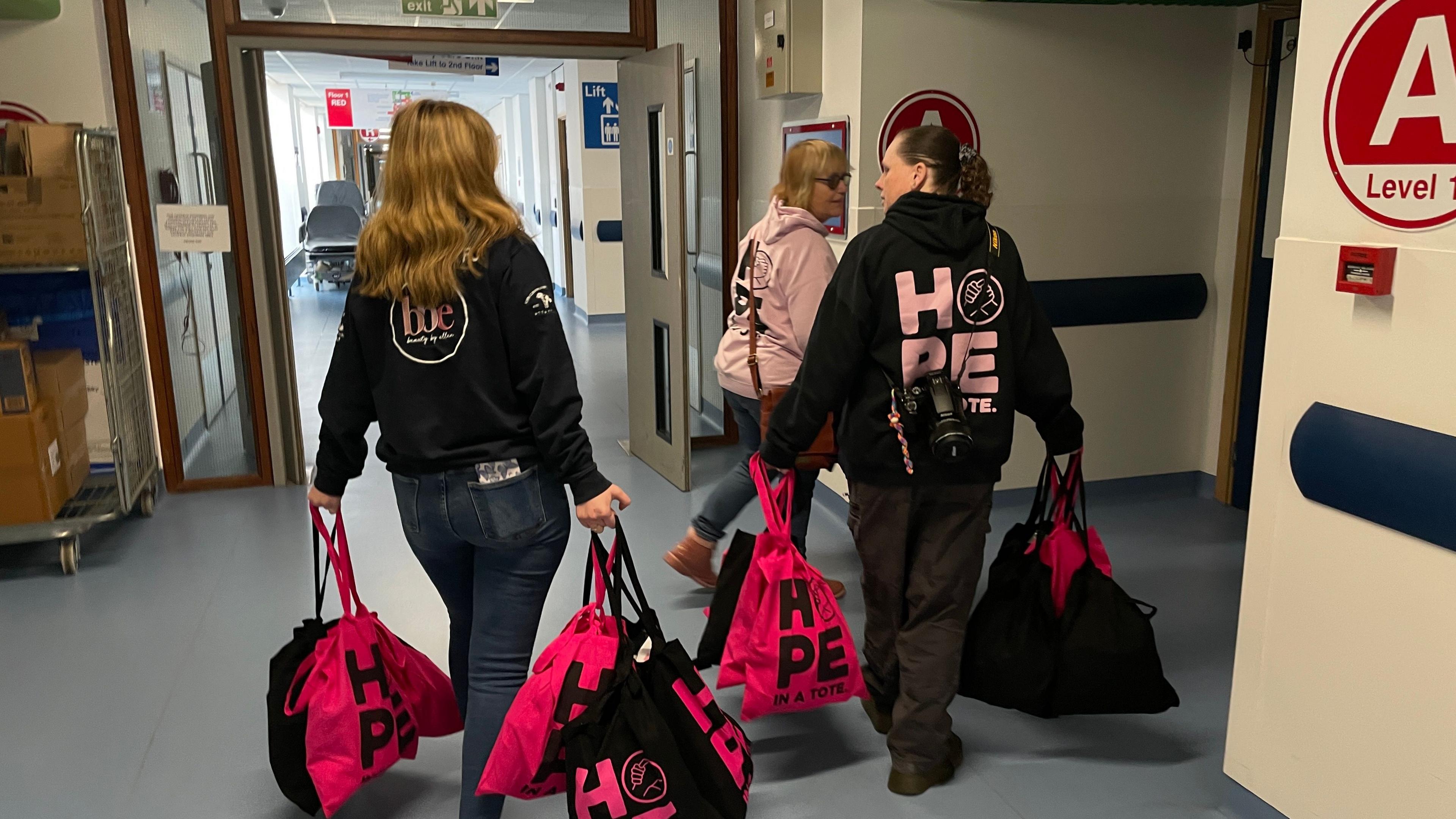 Three women in hoodies which read "hope in a tote" on the back are carrying black and hot pink tote bags down a hospital corridor.
