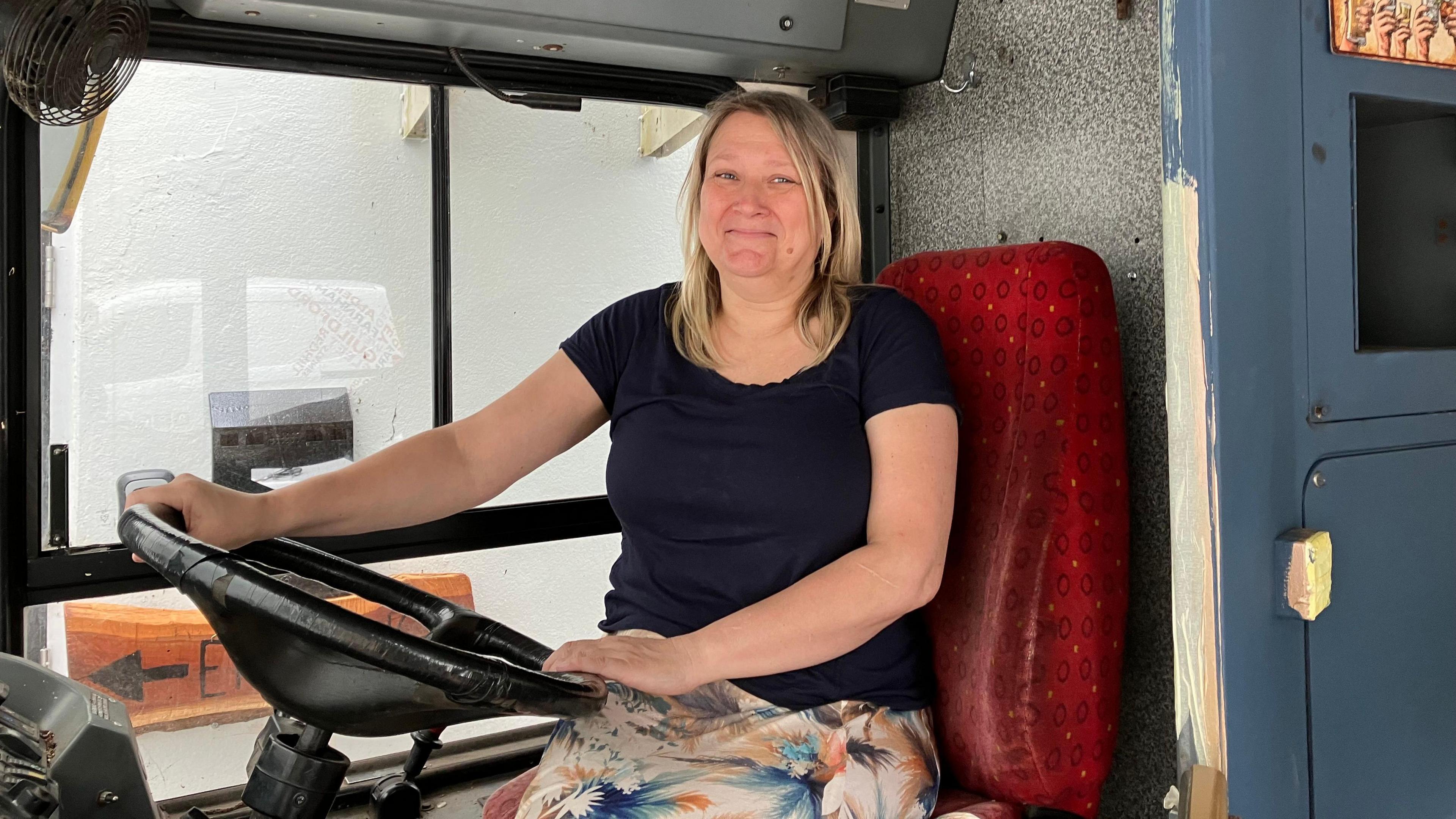 Wendy Salmon sits behind the wheel of the bus, with her hands on it. She is wearing a dark t-shirt and is smiling at the camera. The seat she is sitting on is a covered in a red patterned fabric.