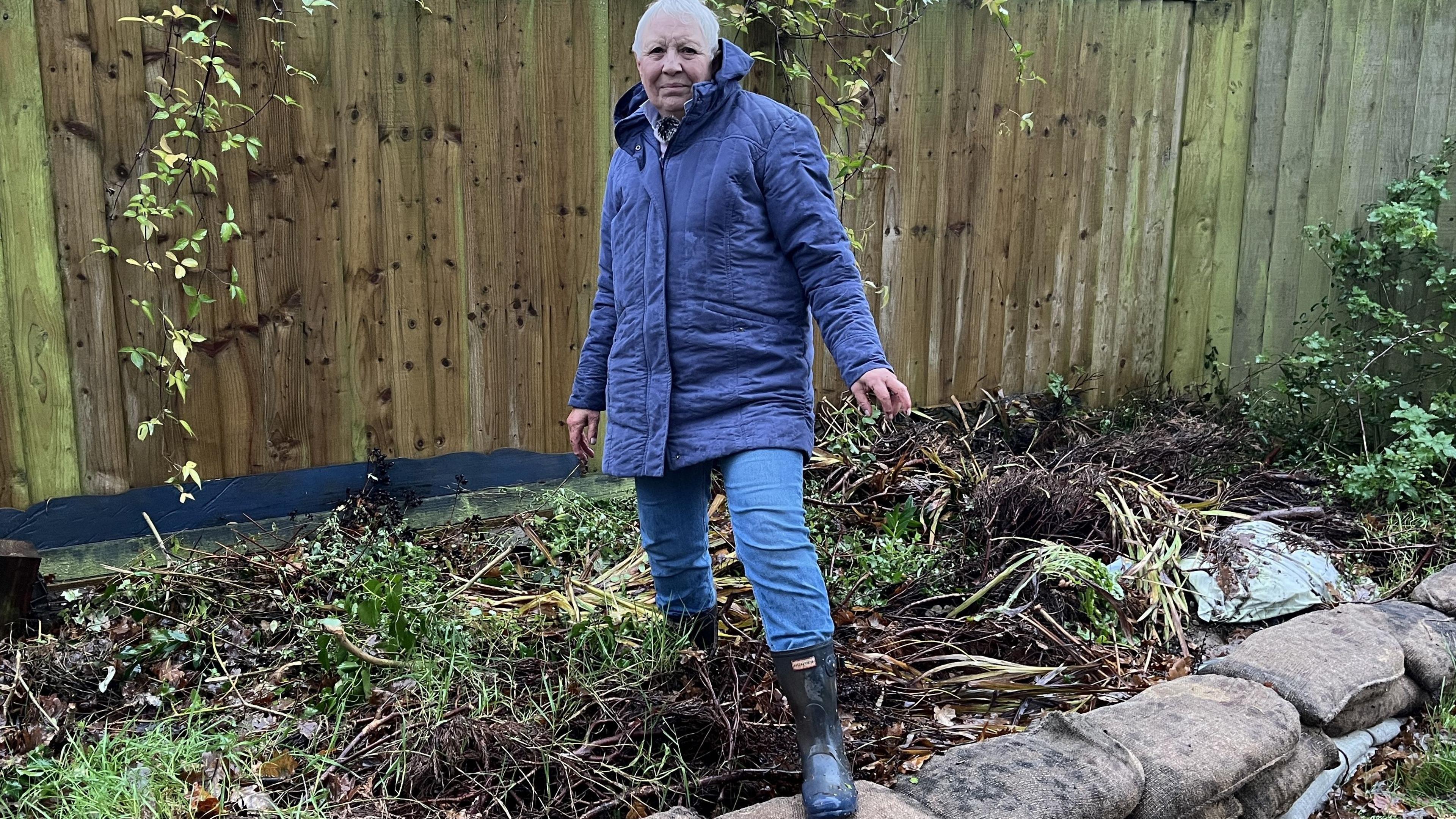 Patricia Clarke standing on the dam she has built, with one foot on a pile of vegetation and the other foot on sandbags