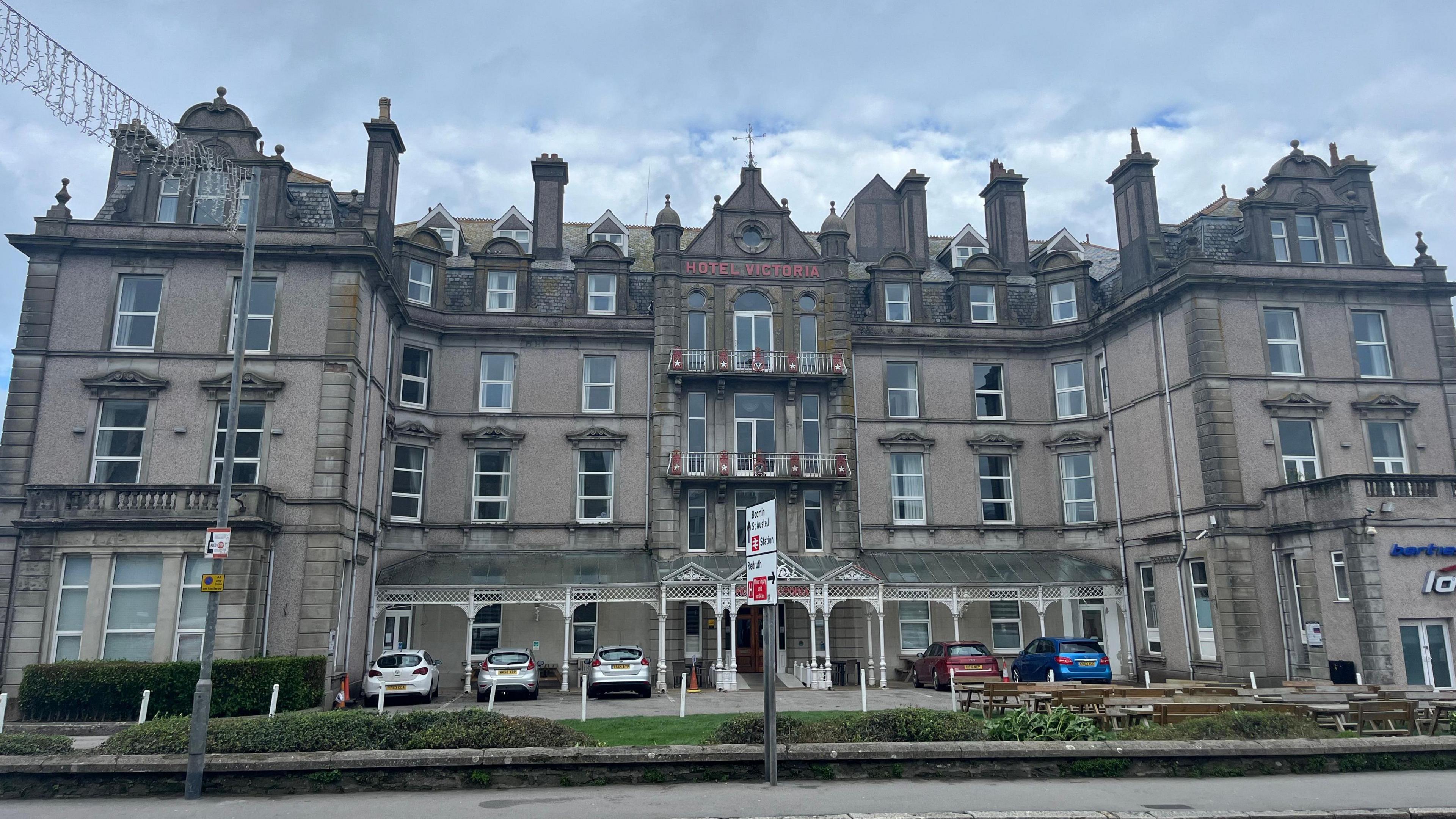 A large gothic hotel with multiple doors and windows is seen with cars in ports at the front. 