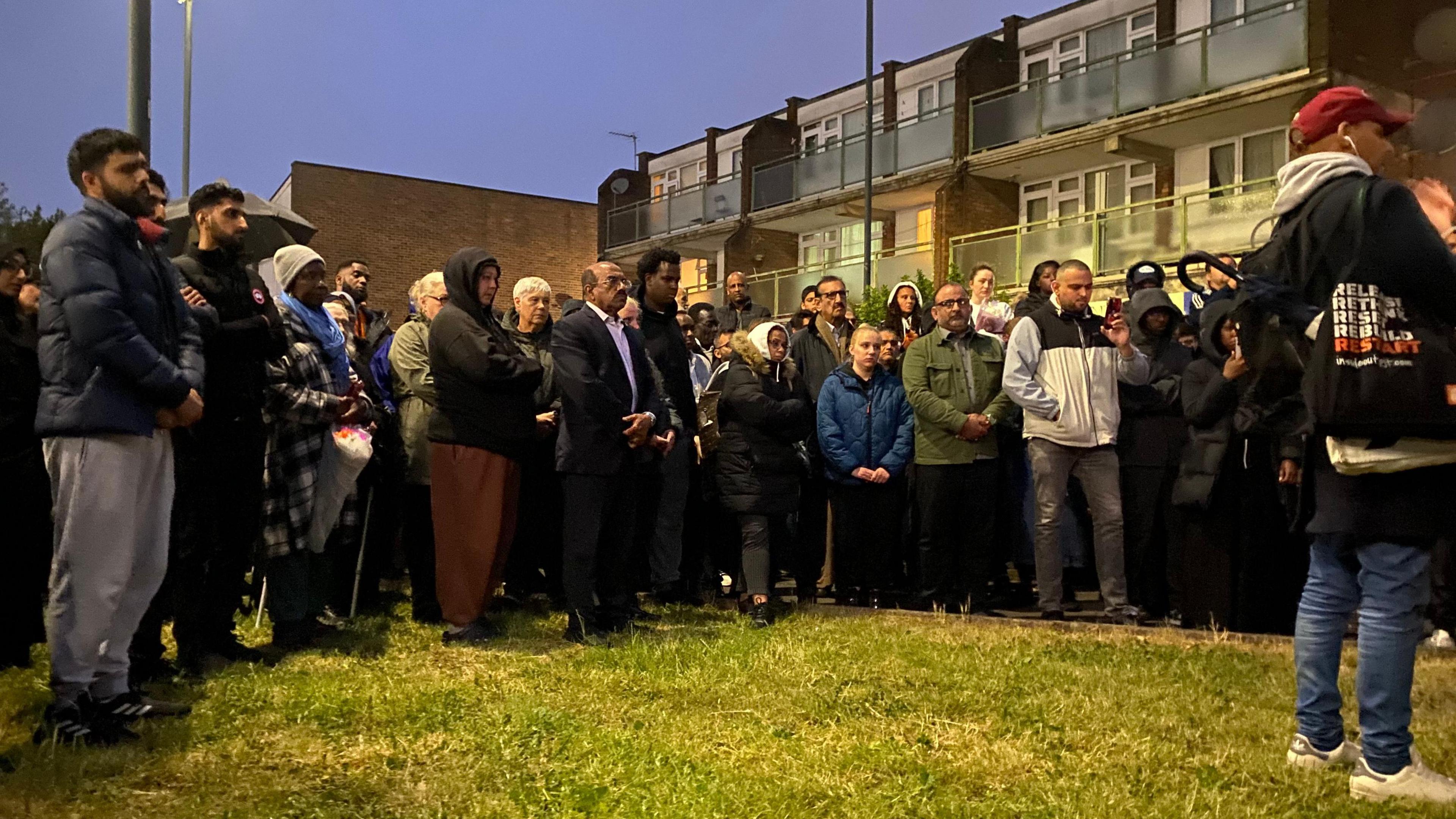 A group of people gather in front of low rise housing at a vigil, in front of a piece of grass. Many have their hands clasped together.