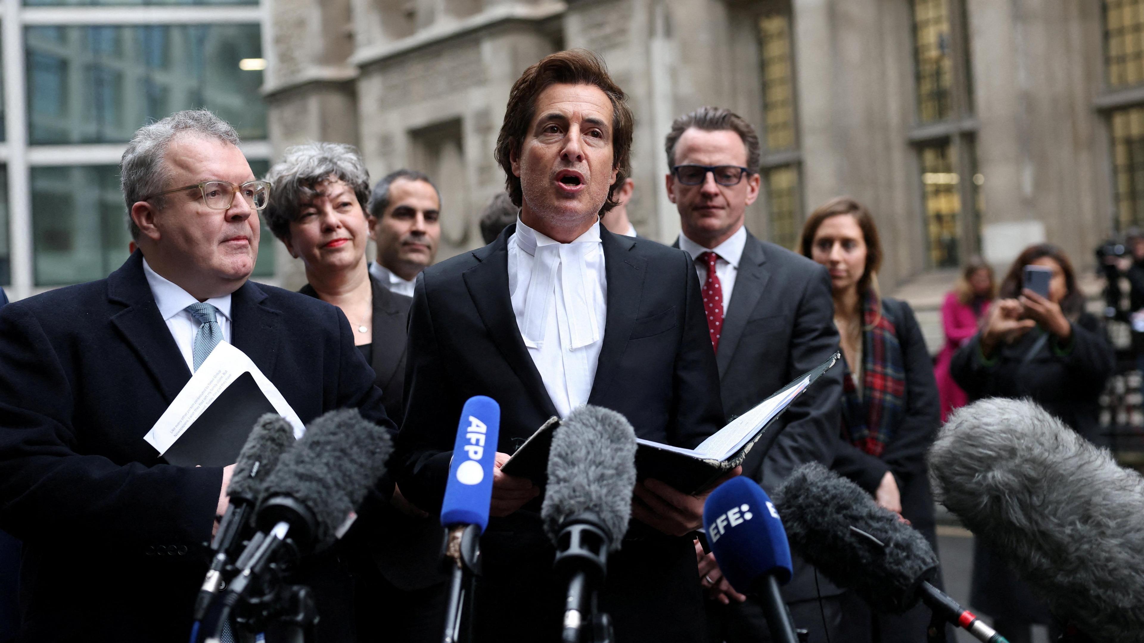 David Sherborne, wearing bands, speaking to reporters at the High Court flanked on the left by Lord Tom Watson, the former Labour MP. Several other people stand behind them and there are several reporters' microphones in the foreground.