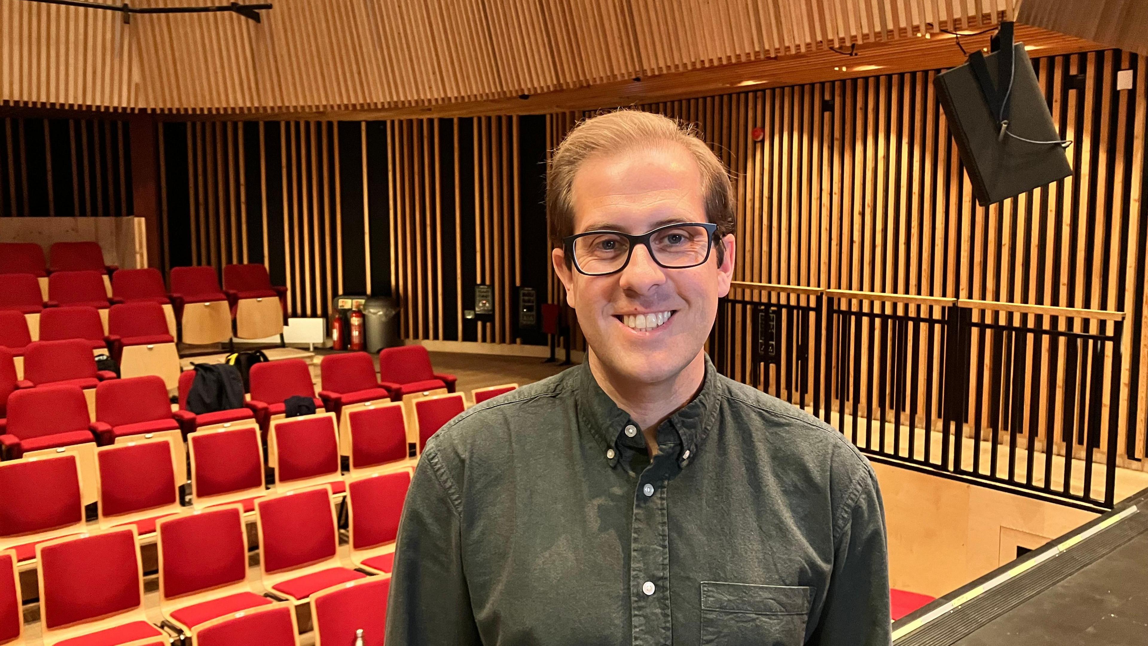 Brendan Paddison, Dean of York St John University's business school, is standing in the university's theatre and has red seats behind him. He is smiling at the camera and is wearing spectacles and a dark shirt.
