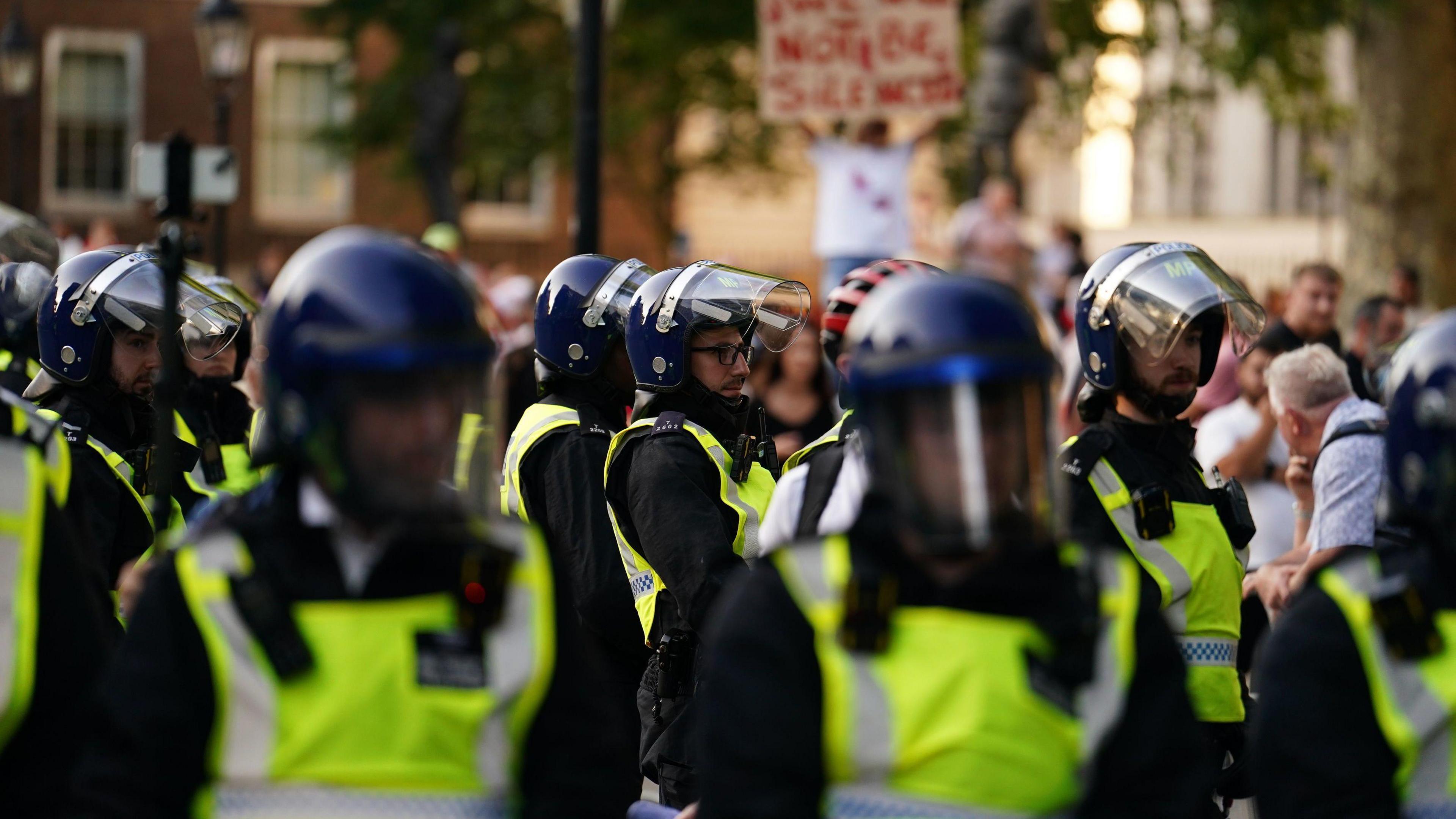 Police officers in riot gear standing in front of protesters. A blurry sign is in the background.