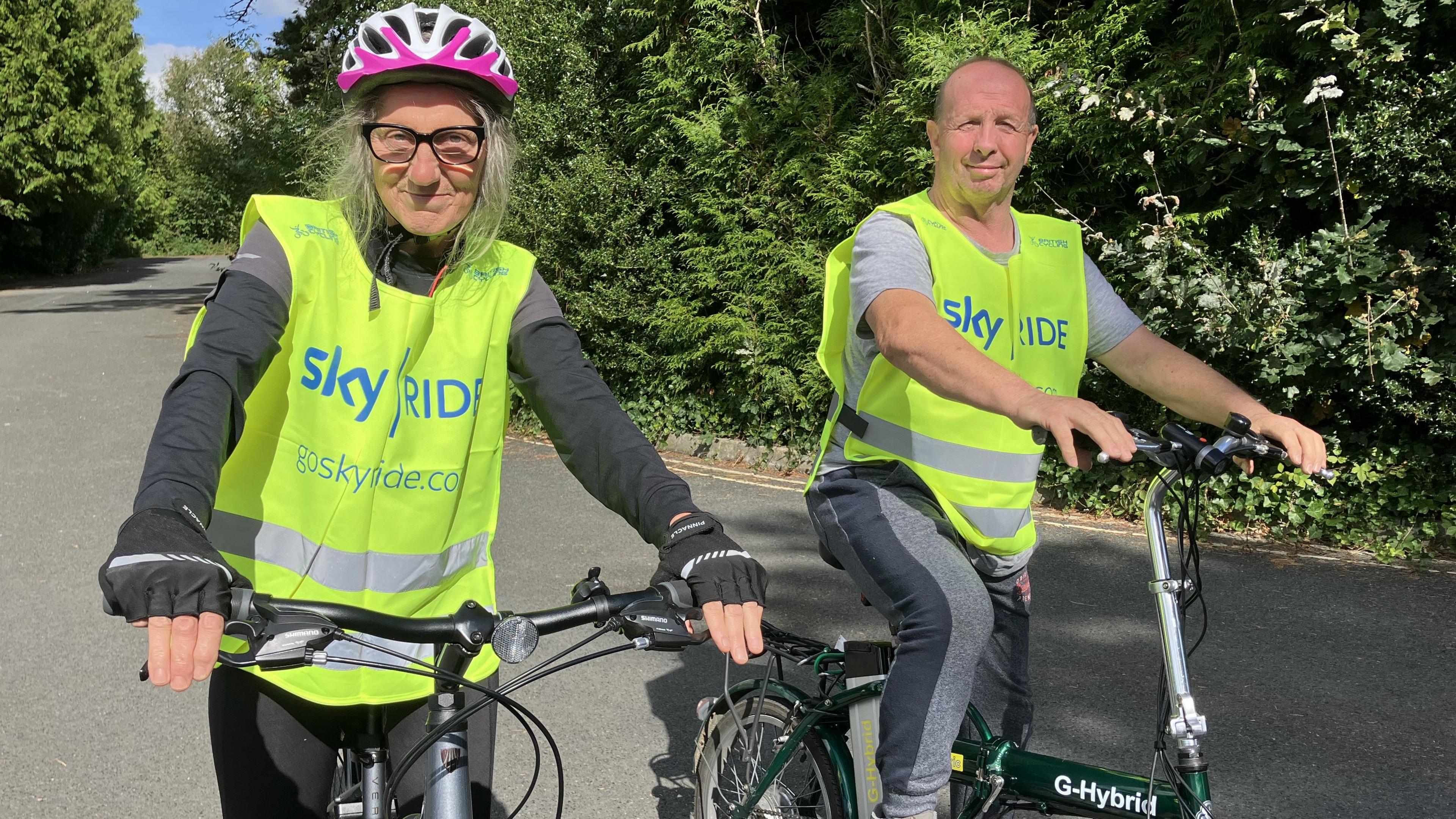 Cycling buddies Lin Harris (left), sitting on a bicycle, wearing a hi-visibility jacket and a riding helmet. Barry Davies is on the right, also sitting on a bike and wearing a hi-visibility jacket