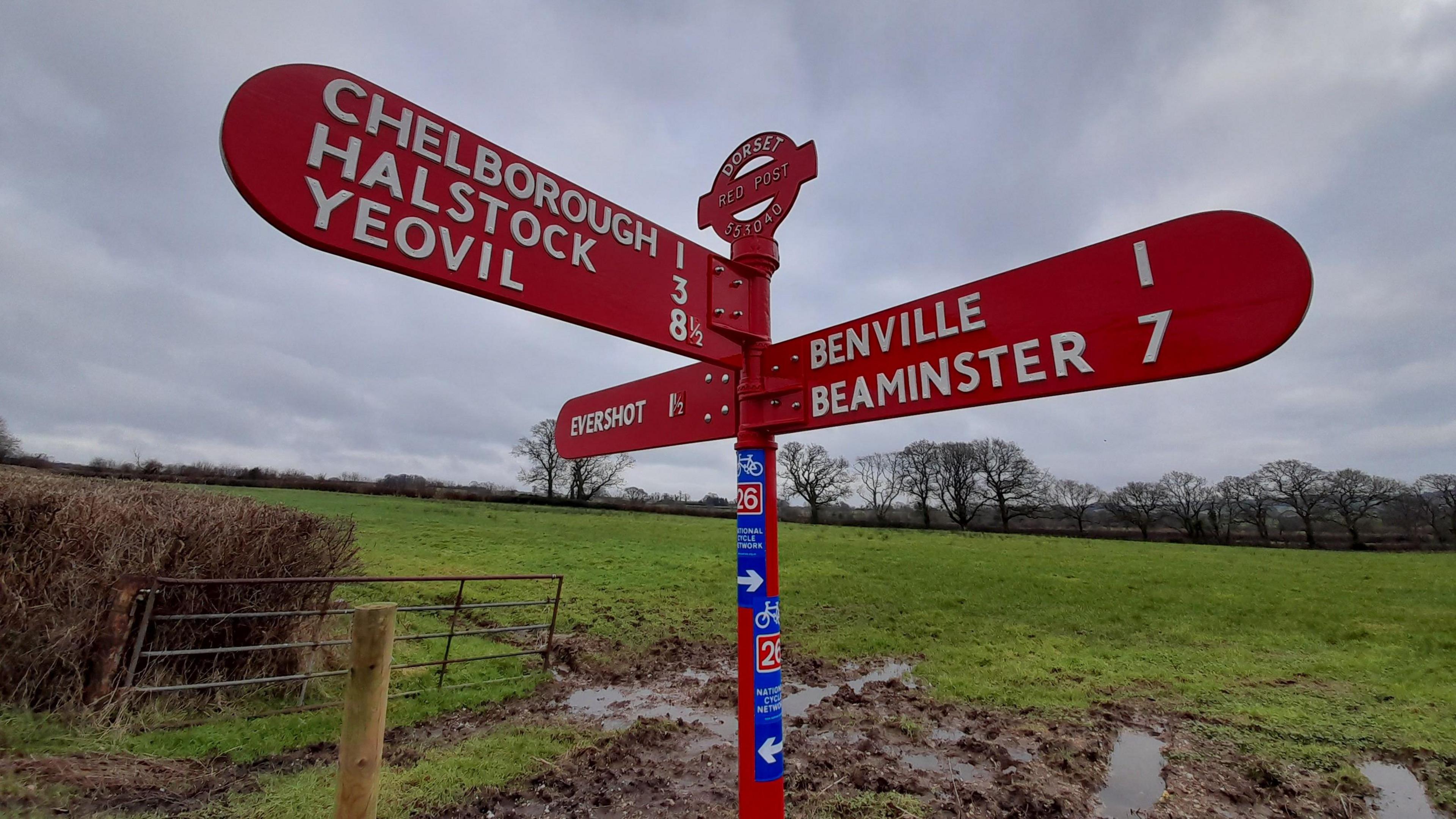 A red, 19th century road sign is positioned in front of a muddy gateway to a field. The sign boards themselves have white lettering and point in three different directions to local villages and towns. National cycle routes are also shown on the main pole. 