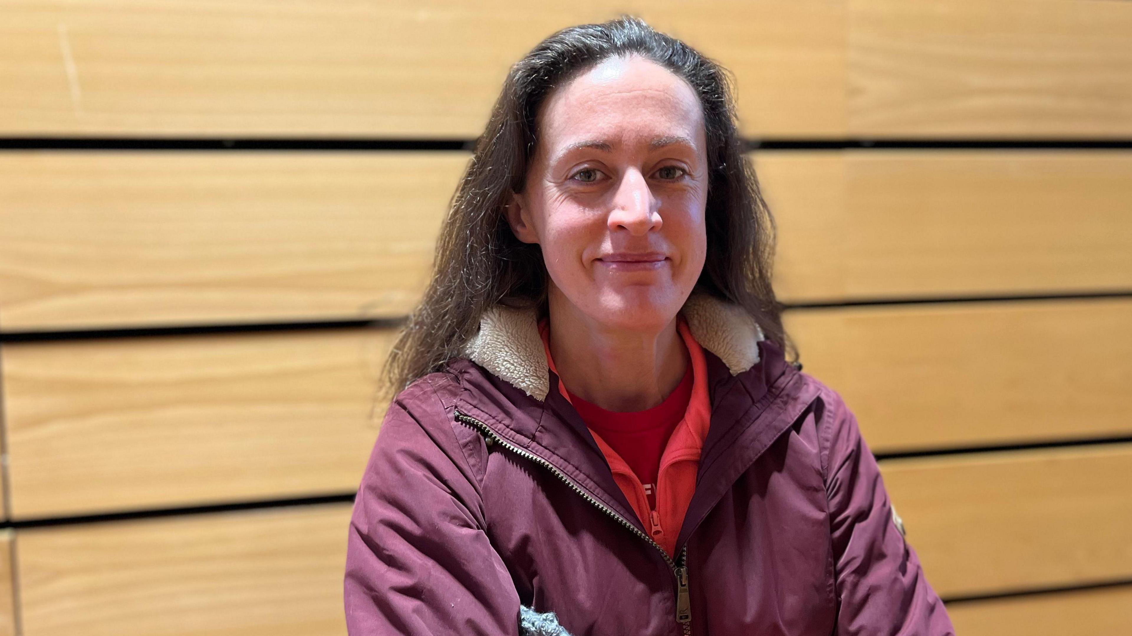 A woman with long dark hair, red top and purple jacket standing in front of a wood-panelled wall