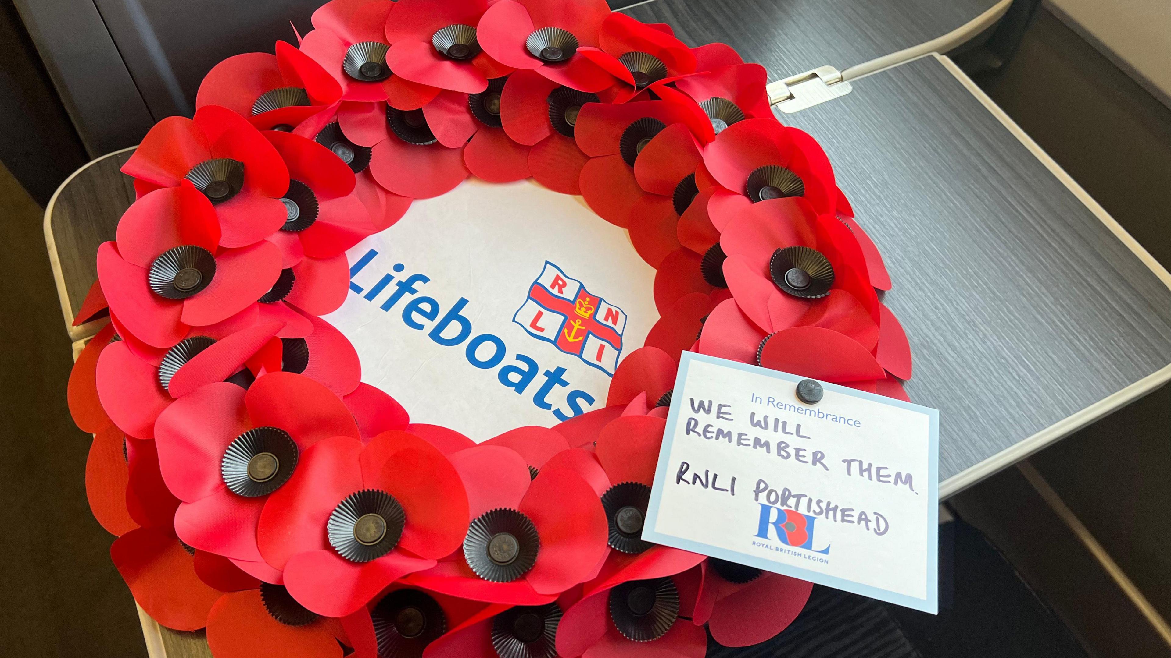 A poppy wreath laid out on a table on a Great Western Railway train. In the middle of the wreath is a white piece of paper with the RNLI logo and the word 'lifeboats' written in blue font. There is a white piece of paper attached to the wreath which says "In remembrance. We will remember them. RNLI Portishead.