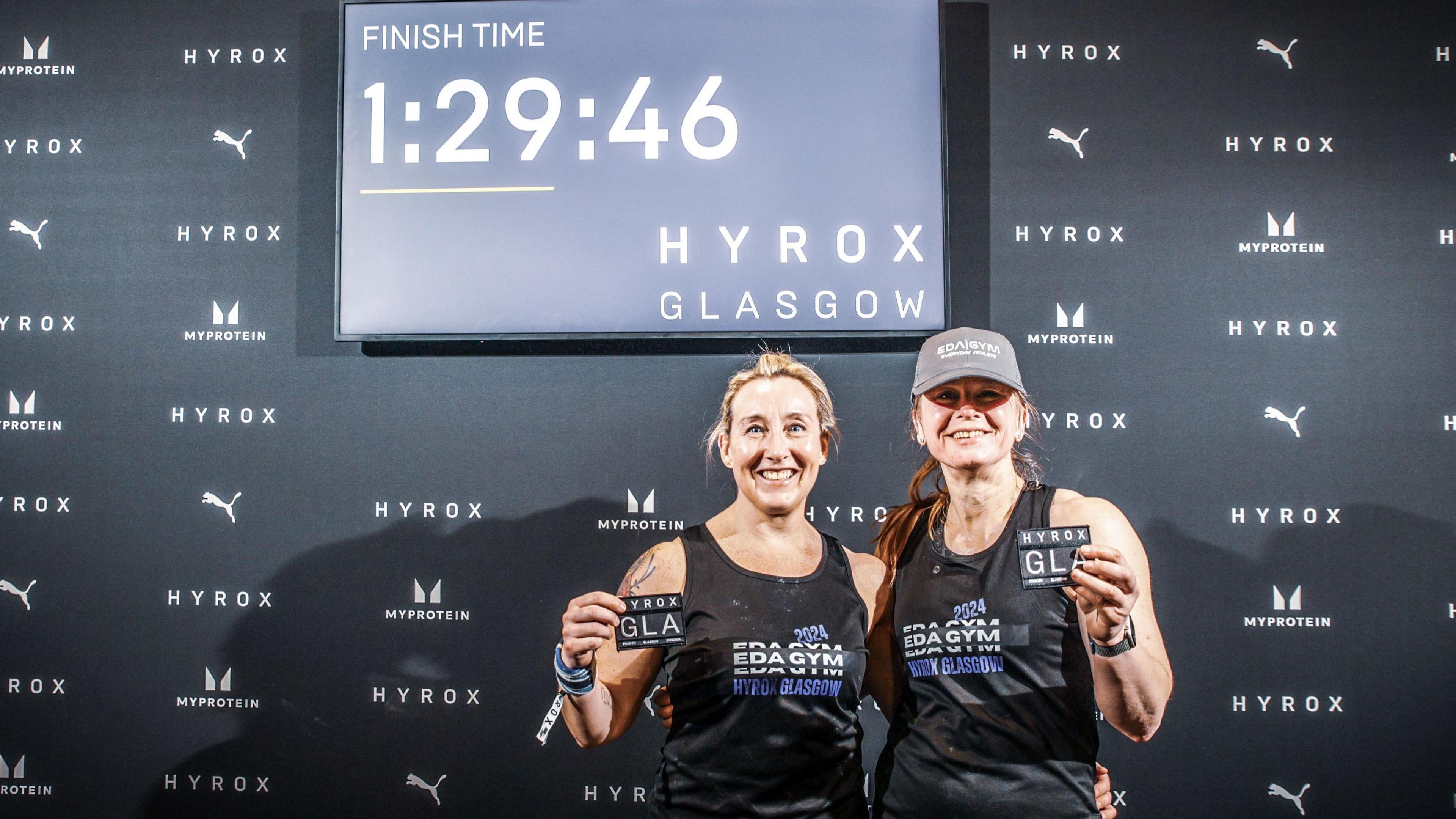 Leigh and Wiki hold up their competition badges under a digital display of their finish time. They both are smiling for the camera.