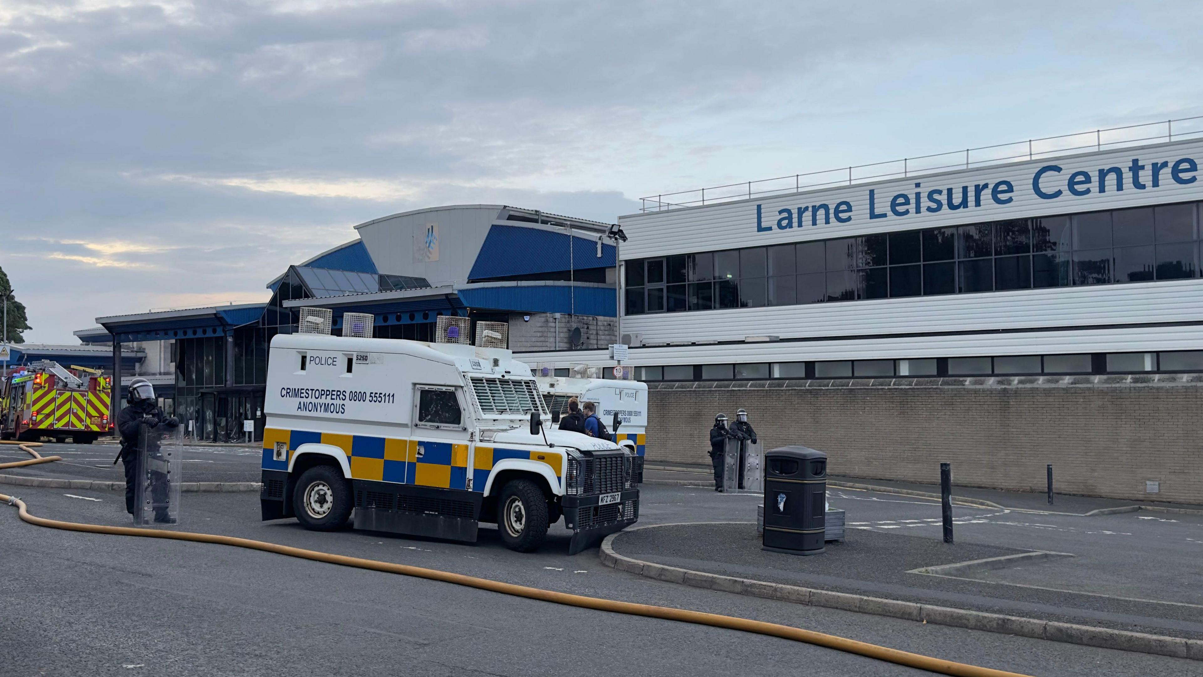 Two white and yellow police vans sit in front of a large grey and black building with the the words 'Larne Leisure Centre' in blue font on it. To the left of the frame a red and yellow fire truck is parked. Three police officer in black riot gear and black helmet hold clear riot shields.