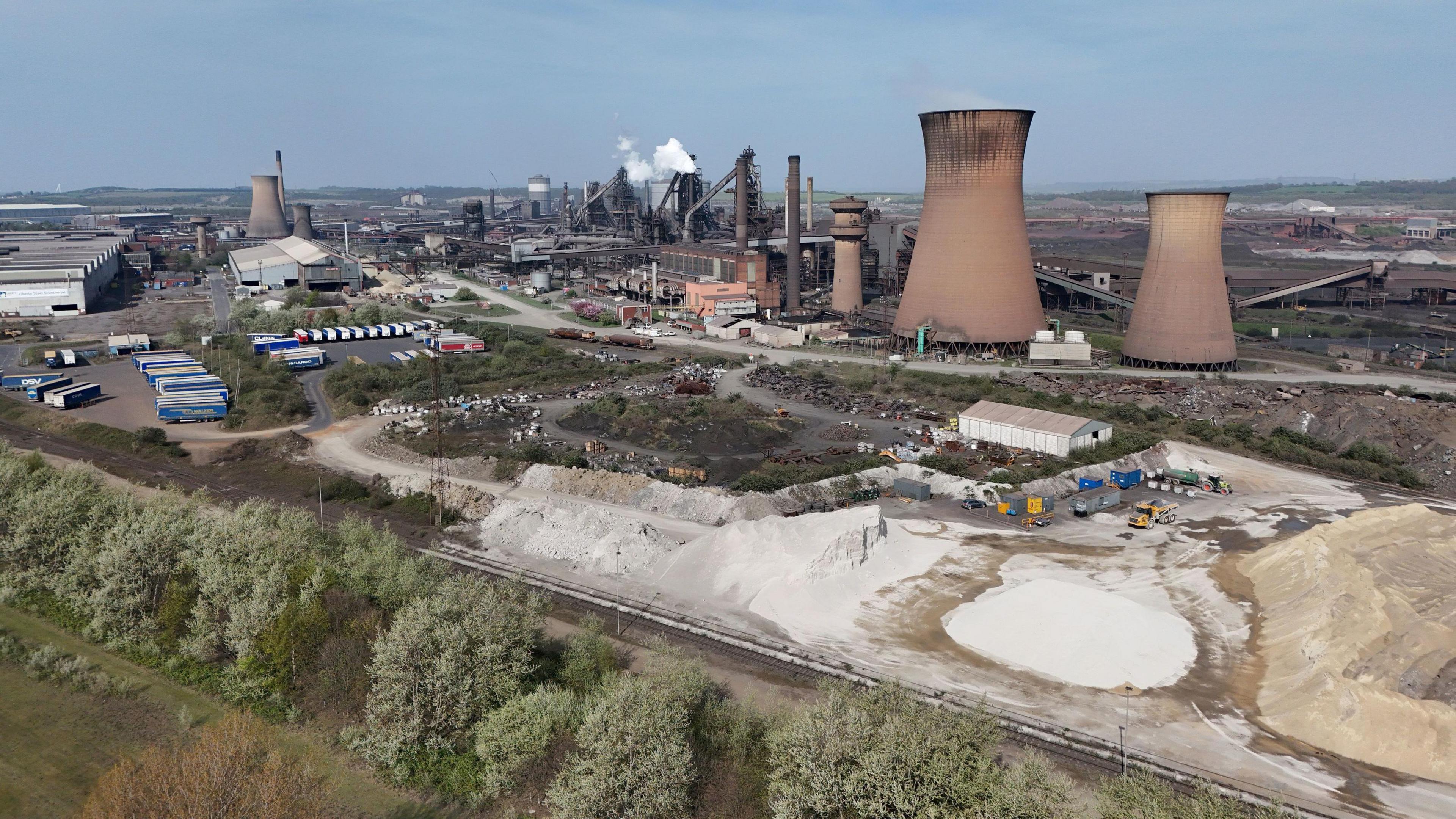 Aerial view of a large steel plant. The site includes cooling towers, large factory buildings, and piles of raw materials that appears to be sand. Machinery and storage containers are scattered throughout. Smoke rises from some structures, Trees and greenery border the bottom of the site.