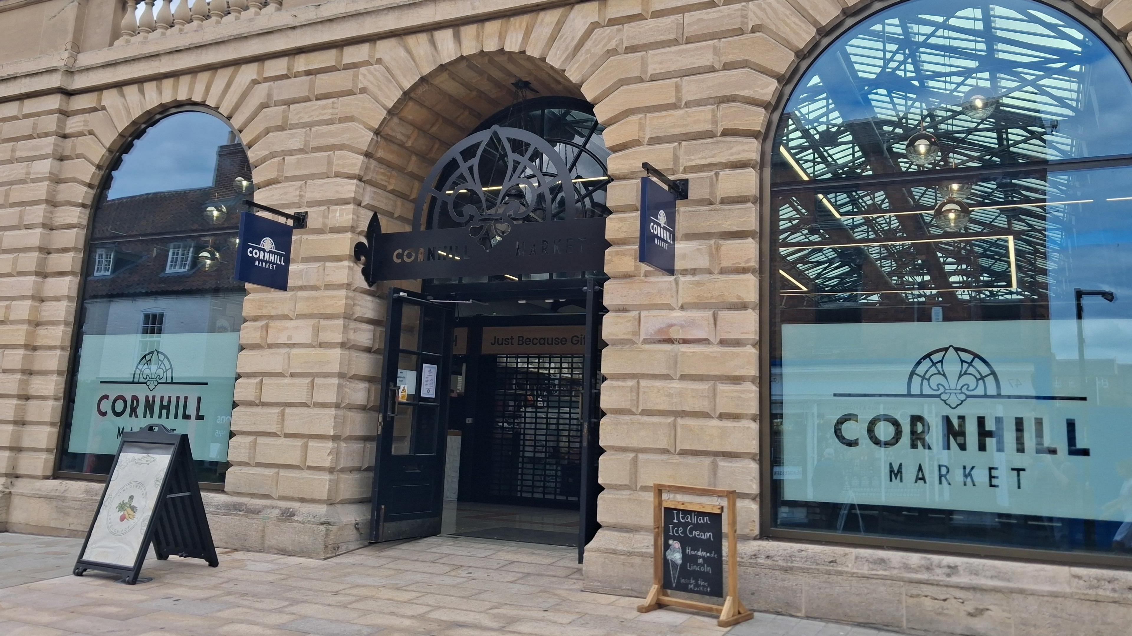 A sand-coloured brick building with two arched glass windows and an arched entrance. There are two freestanding signs outside. On the windows it says "Cornhill Market".
