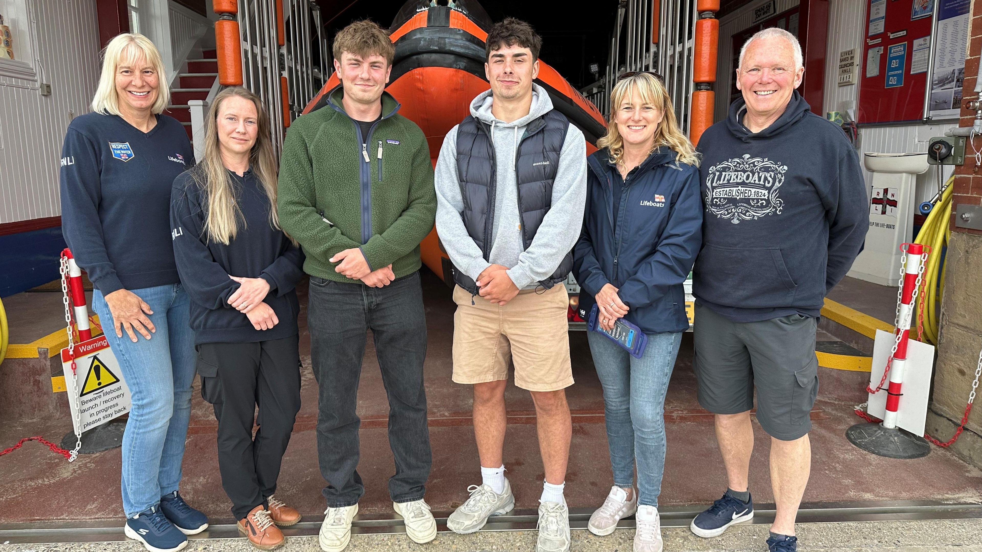 The two brothers stand with four RNLI volunteers, three women and a man who are wearing navy RNLI jackets. Behind them is the lifeboat.
